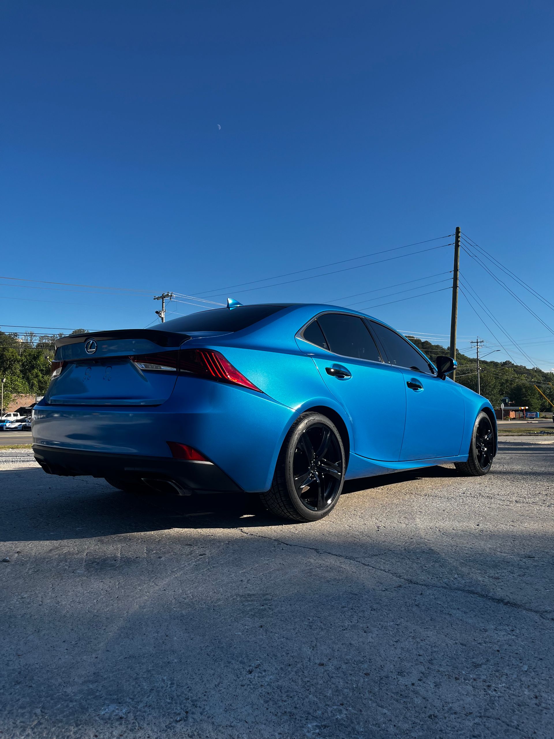 A bright blue sedan with tinted windows and black rims, parked on a gravel lot under a clear blue sky.