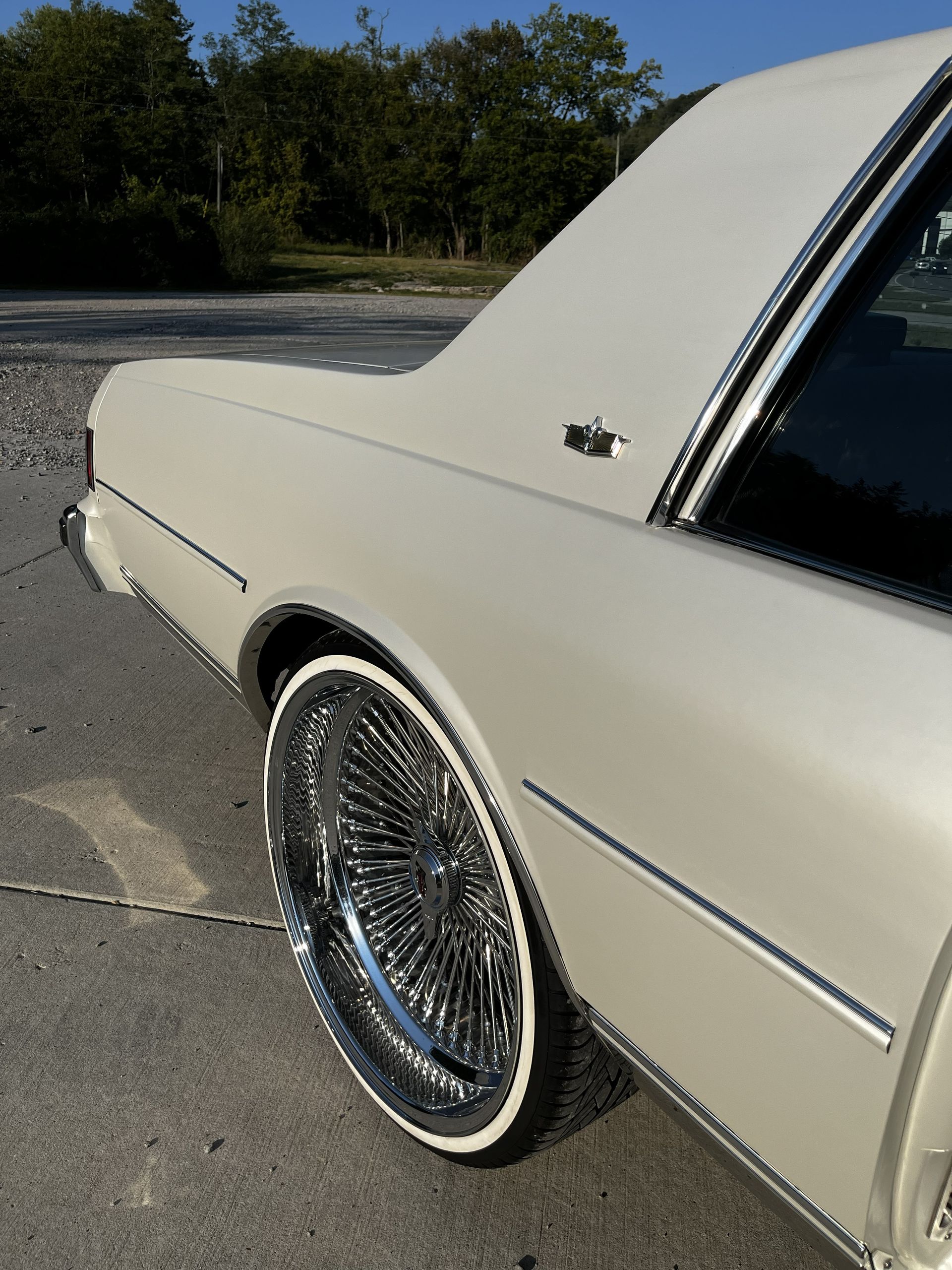 A beige vintage sedan featuring large chrome wire-spoke wheels with thin whitewall tires parked on asphalt.