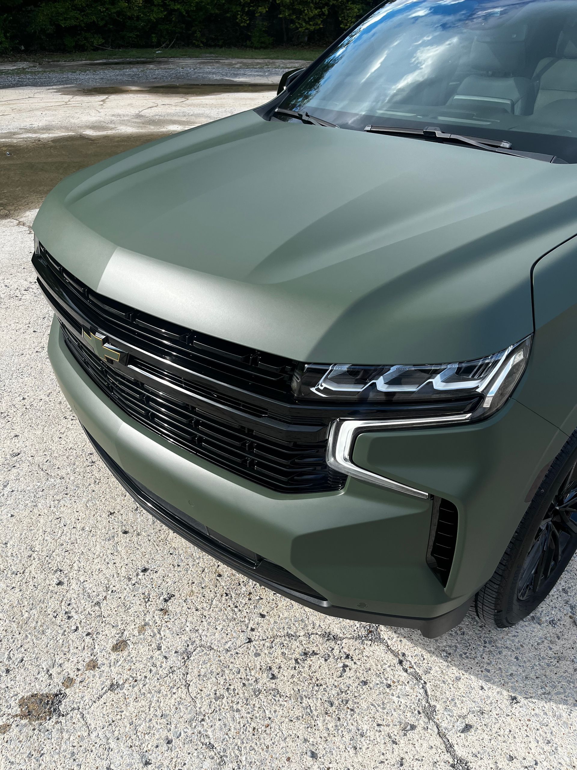 A matte olive green Chevrolet Tahoe SUV parked on a light-colored gravel lot, viewed from the front passenger side.