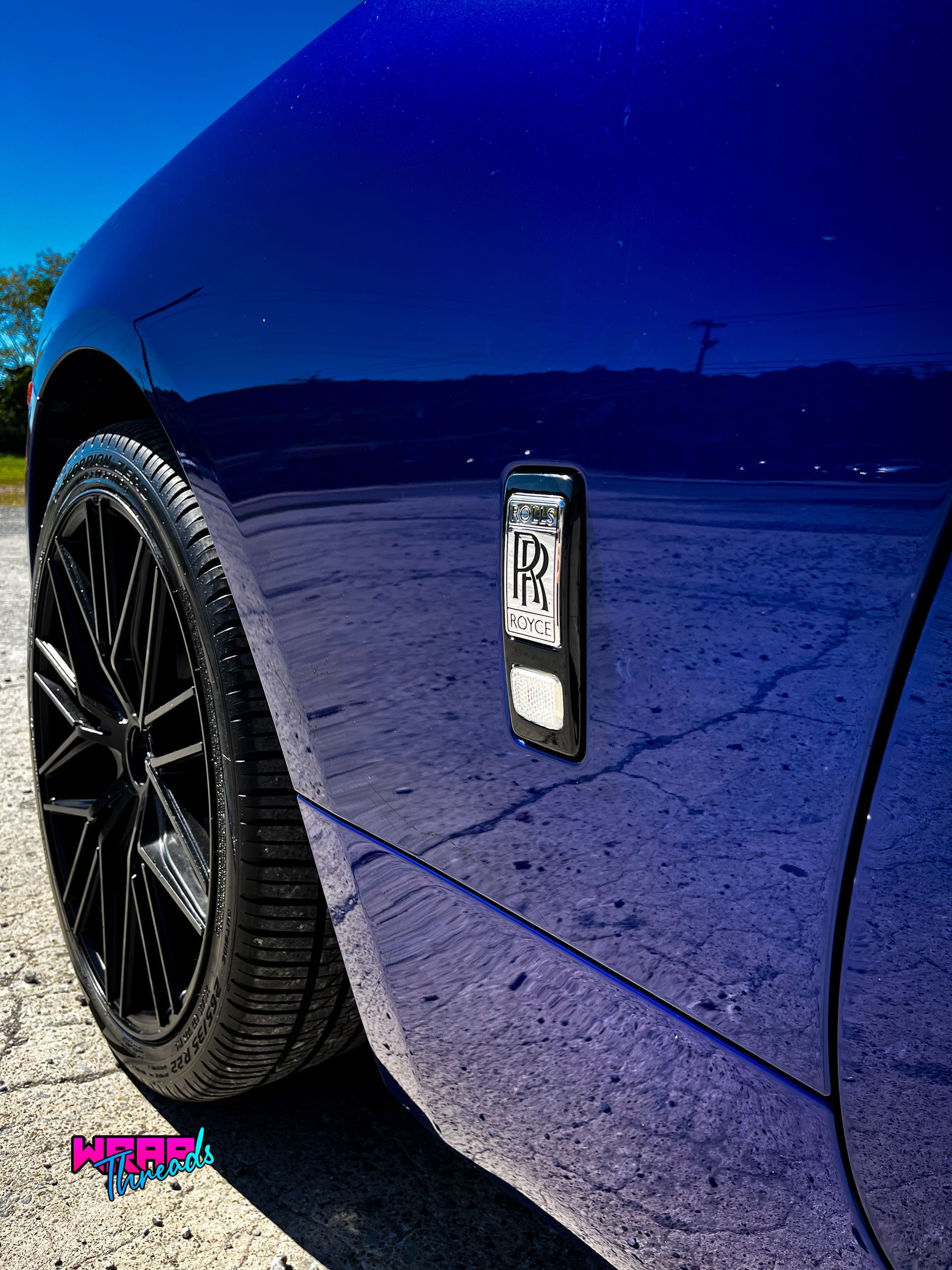 Close-up of a blue Rolls-Royce car body with a prominent, dark-finished badge and black alloy wheels on a paved surface.