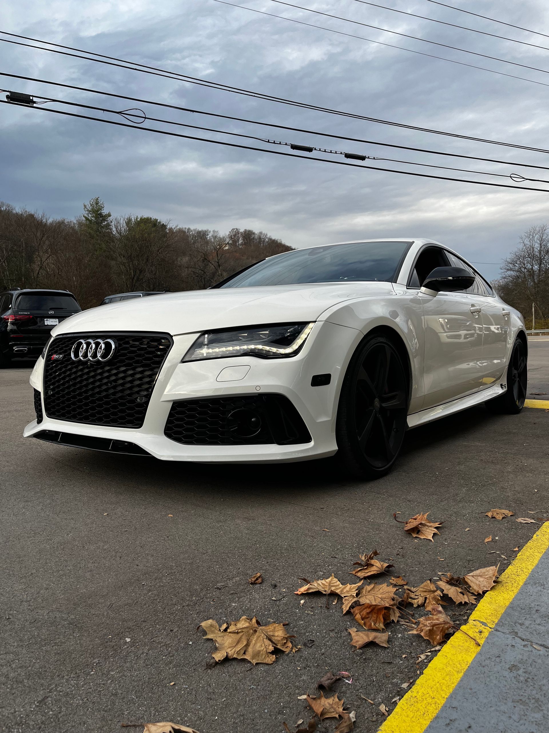A white Audi RS7 parked on a paved road with scattered autumn leaves and overhead power lines under a cloudy sky.