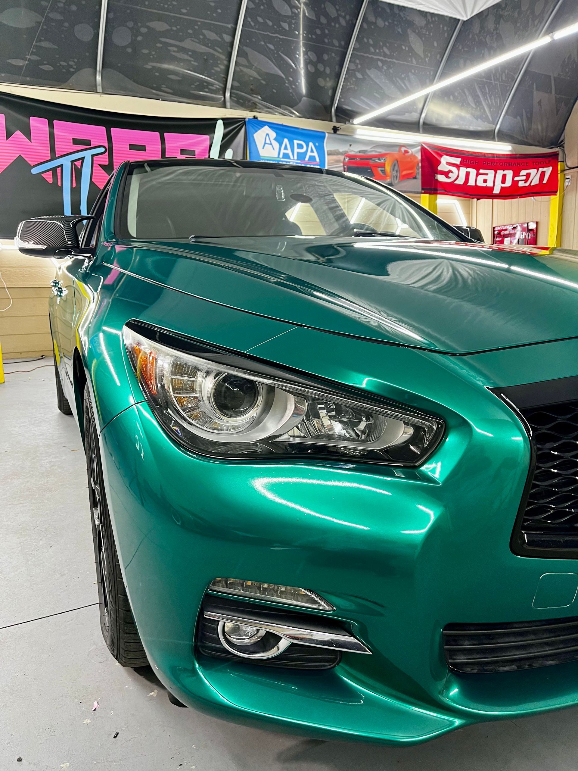 A metallic emerald green Infiniti sedan parked inside a garage workshop with Snap-on signage in the background.