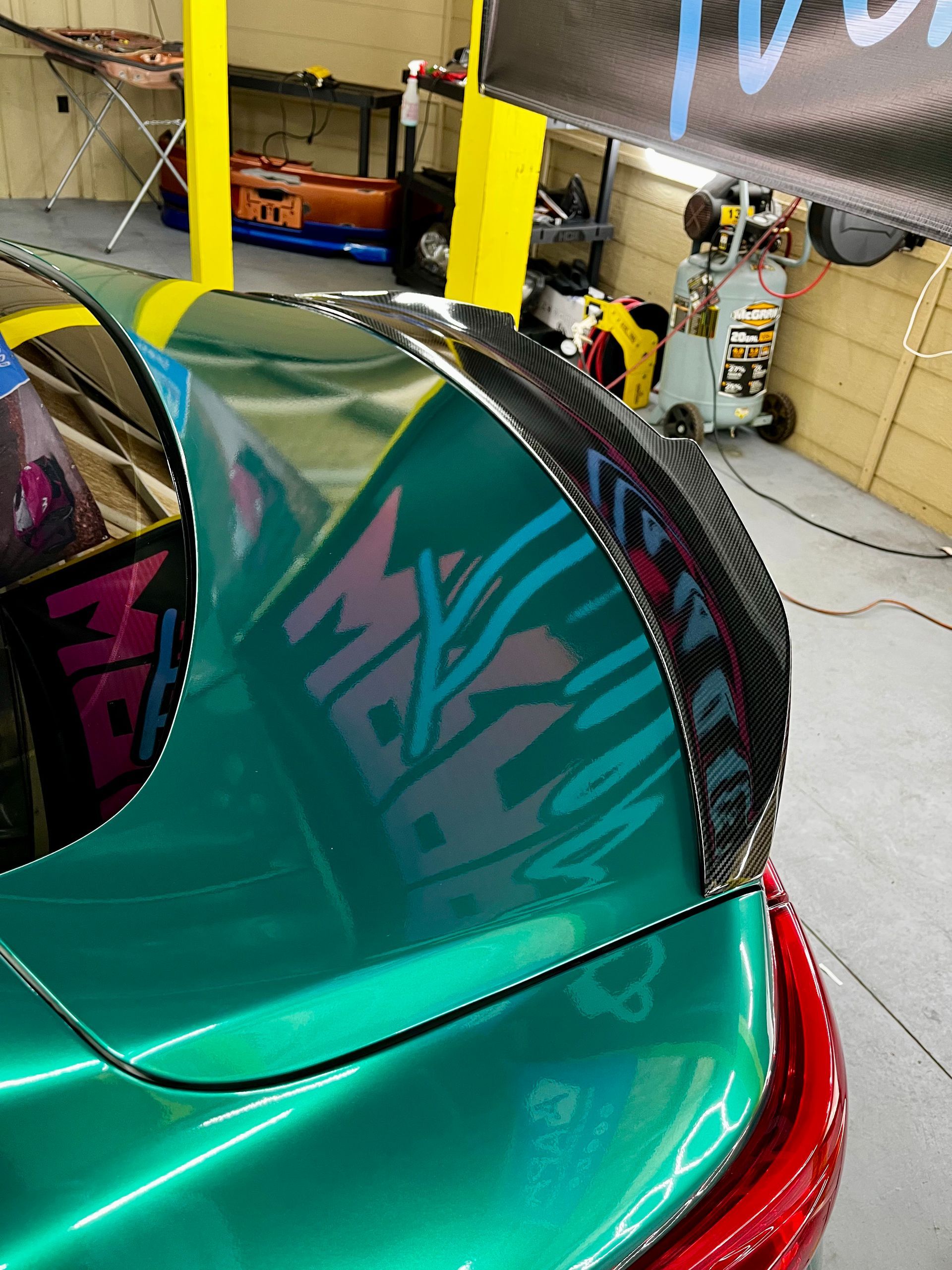 A close-up of a metallic teal car trunk featuring a curved carbon fiber spoiler in an automotive shop setting.