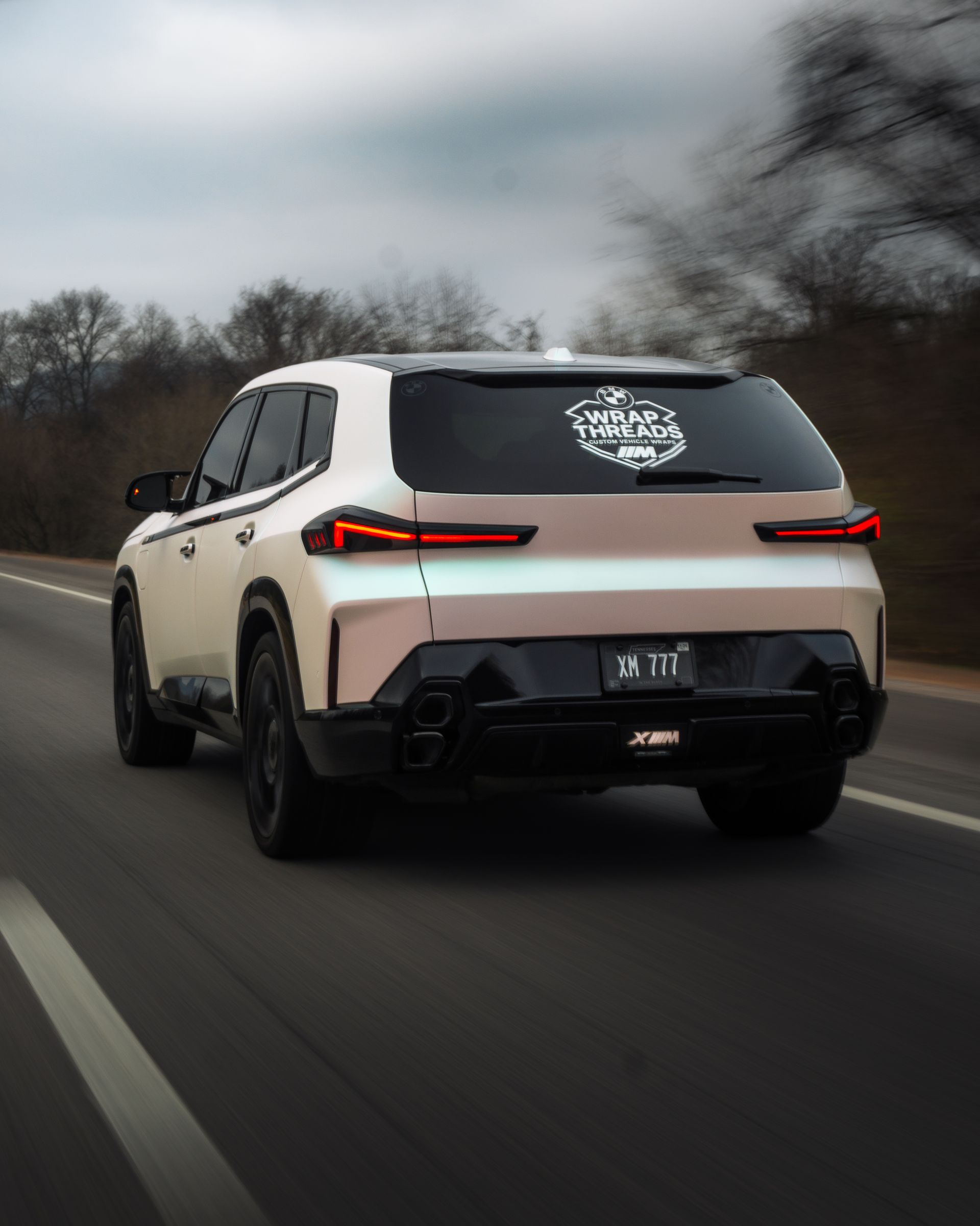 A white BMW XM driving on a road, captured from the rear, with a logo sticker on the back window.