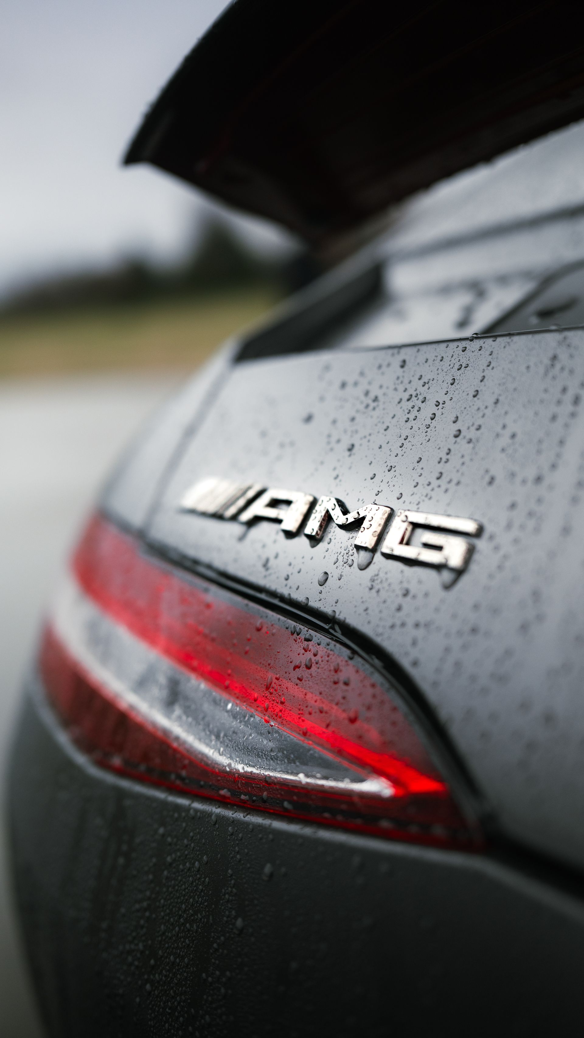 Close-up of a matte black Mercedes-AMG car trunk with a chrome emblem, featuring a red taillight and raindrops on the surface.