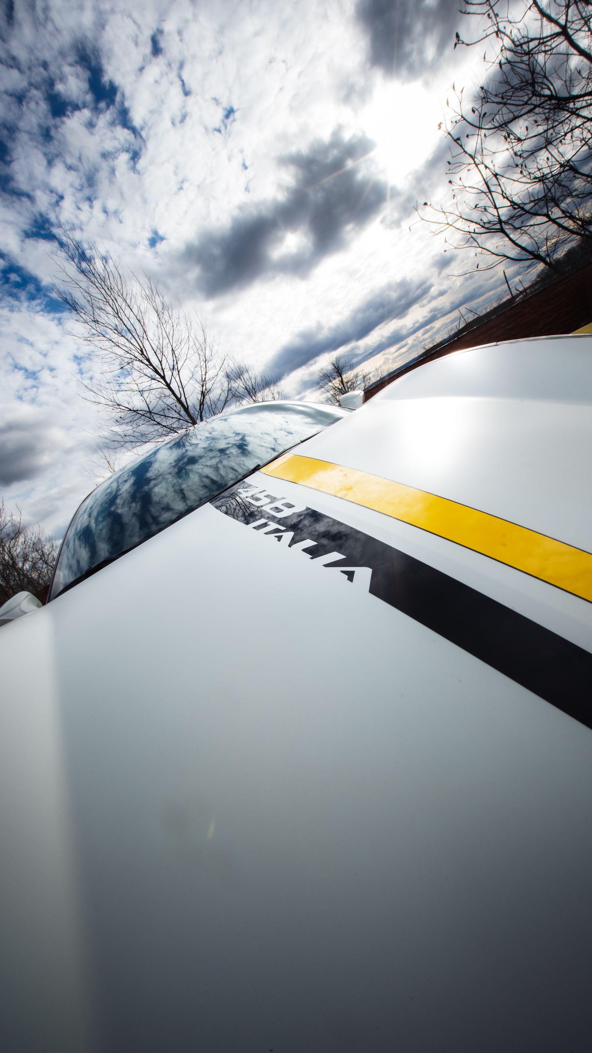 A low-angle view of a white car hood featuring a black and yellow racing stripe, set against a cloudy blue sky.