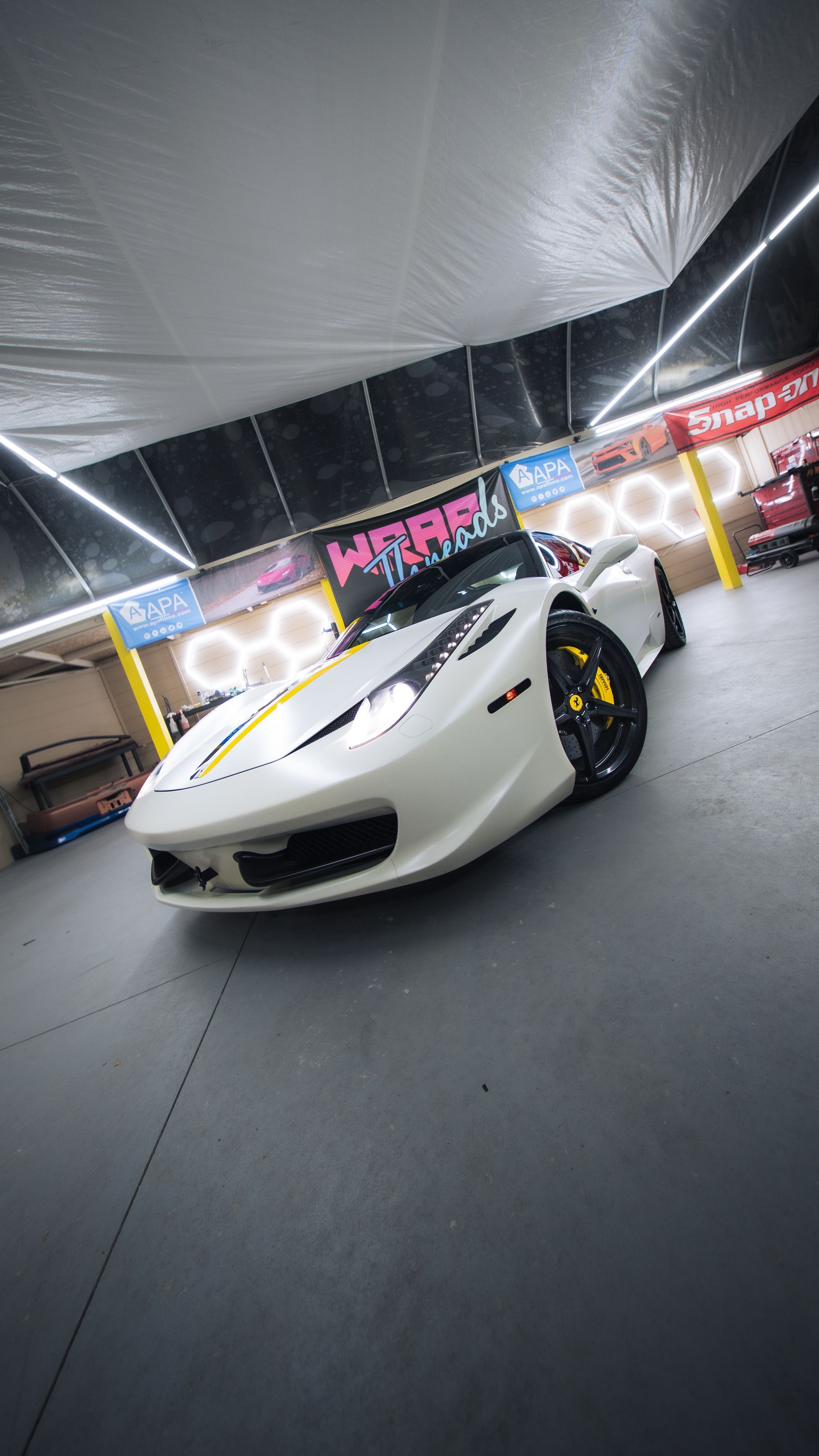 A white Ferrari with yellow brake calipers parked in a garage with neon-lit wall decorations.