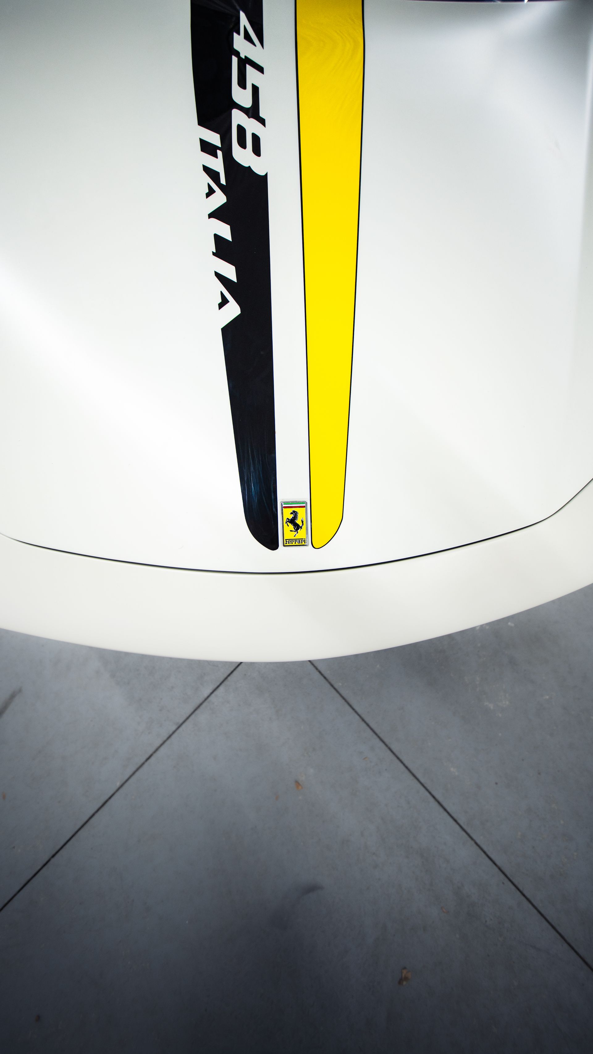 A white hood of a Ferrari 458 Italia with a vertical black and yellow racing stripe and a small Ferrari logo emblem.