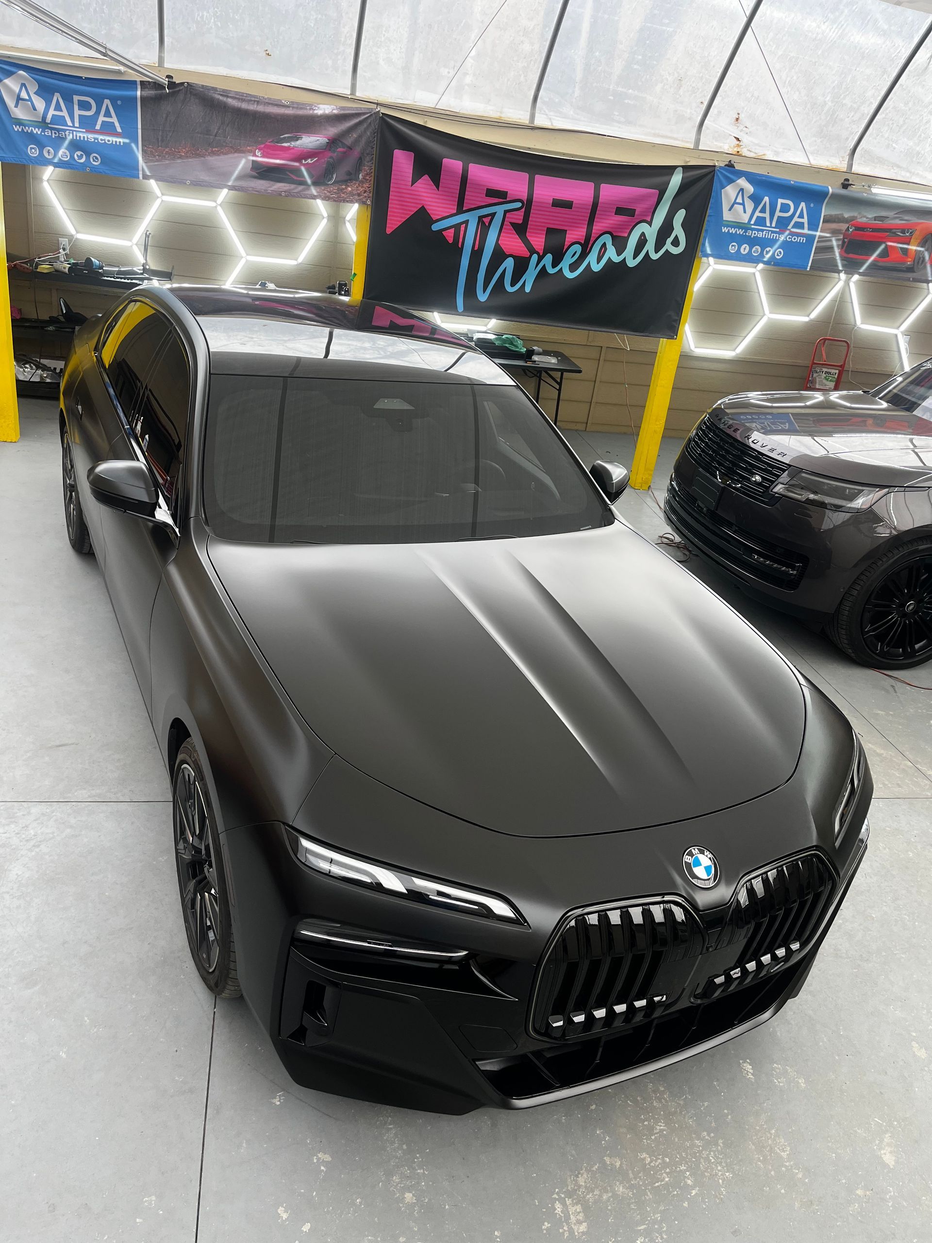 Matte black BMW 7 Series parked in a vehicle wrapping shop with professional lighting and branded signage.