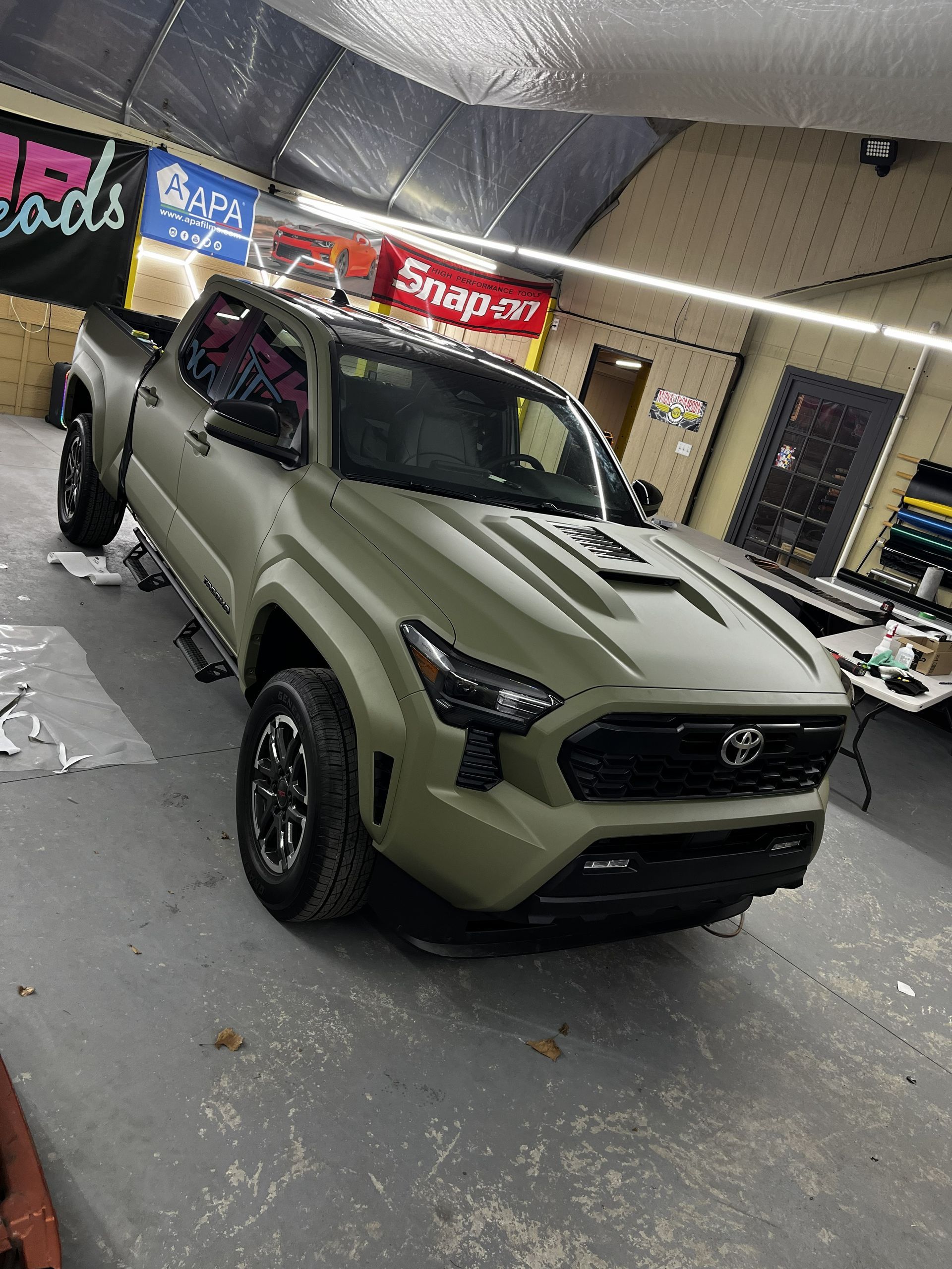 An olive green Toyota Tacoma truck sits inside a garage, currently undergoing a vehicle wrap process.
