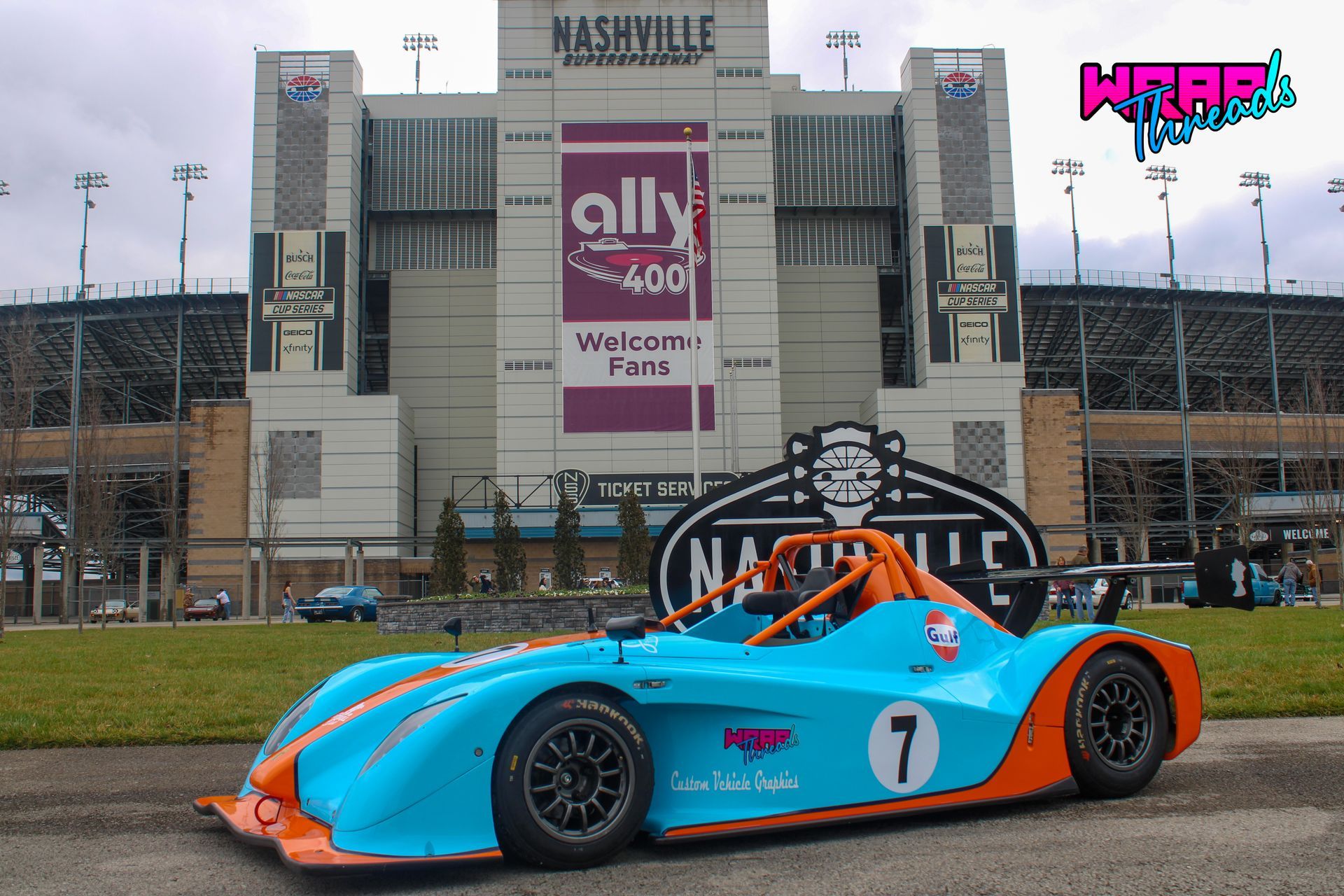 A turquoise and orange race car with the number 7 parked in front of the Nashville Superspeedway stadium.