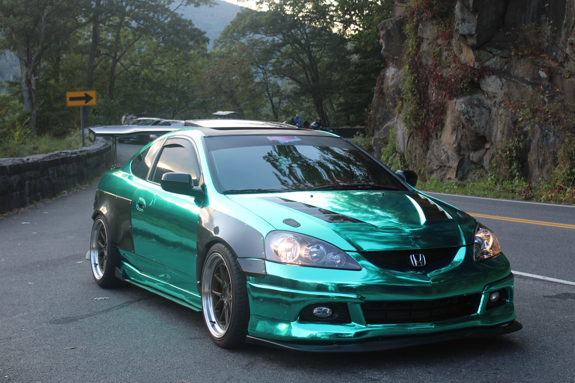 A metallic green Acura RSX with wide fender flares driving on a scenic, winding mountain road.
