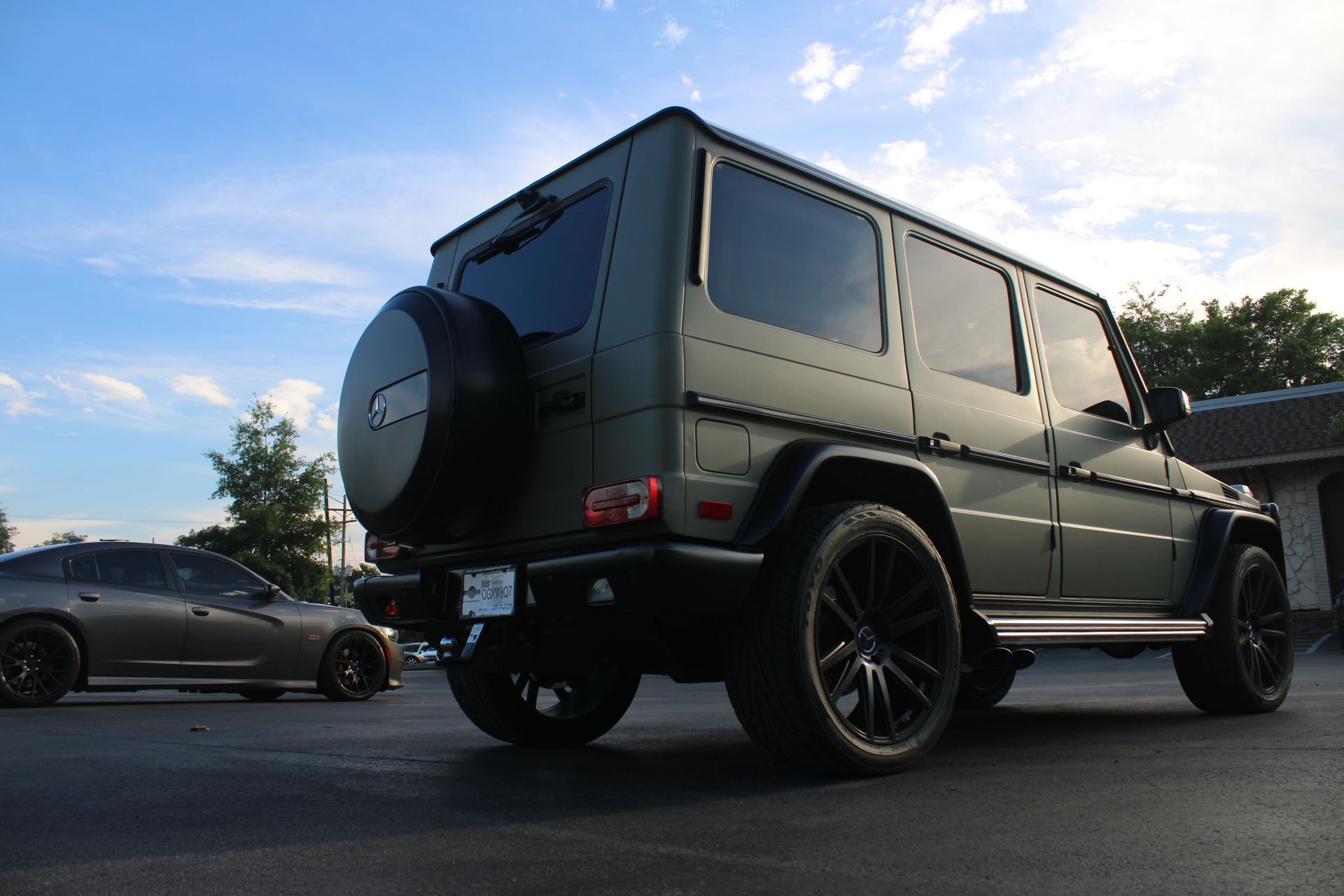 A matte olive-green Mercedes-Benz G-Wagon parked outdoors, with a dark-colored sedan partially visible in the background.
