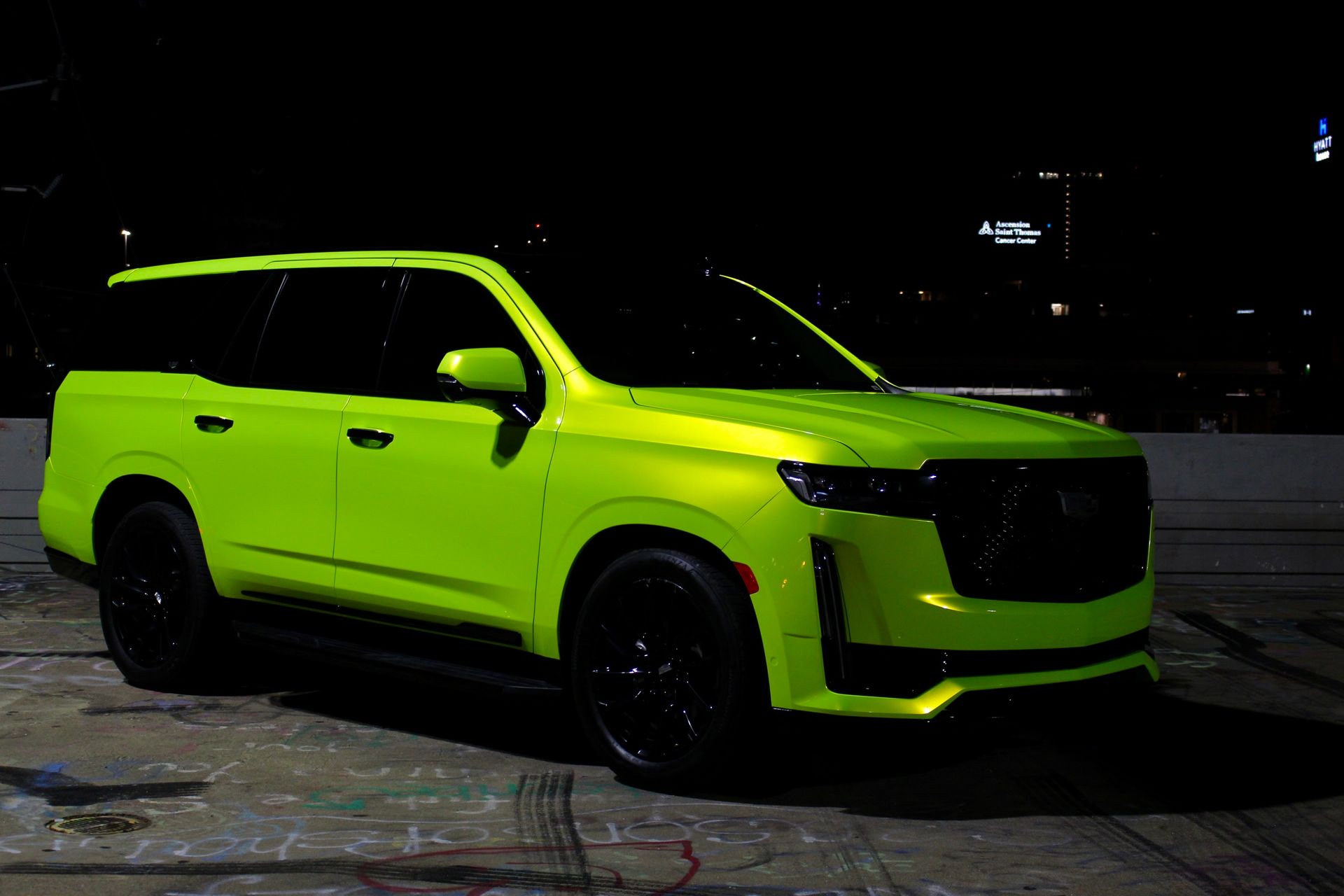 A bright neon-green Cadillac Escalade SUV parked in a dark, dimly lit outdoor setting at night.