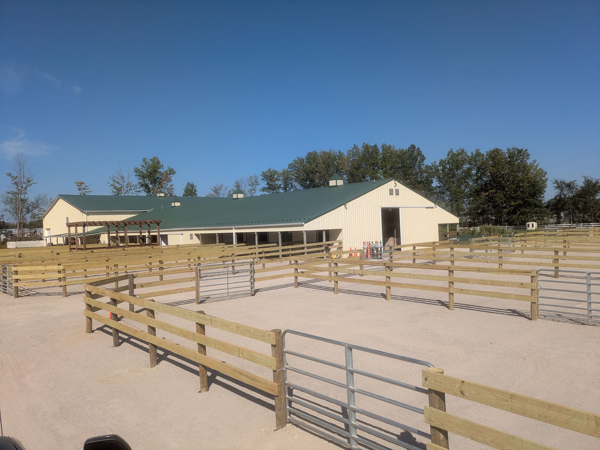 Equestrian facility with tan barn, green roof, tan fencing, and multiple paddocks under a blue sky.