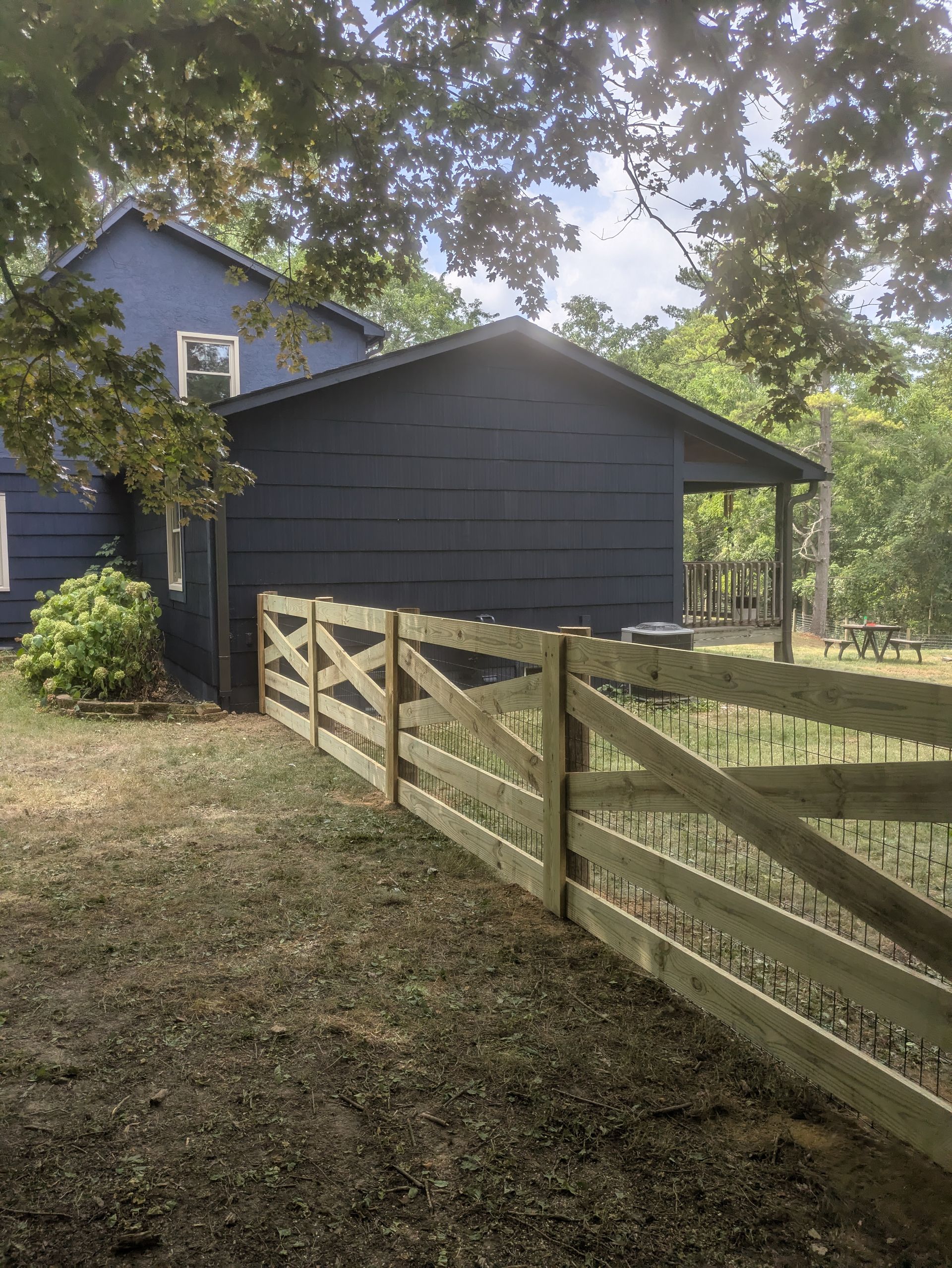 Blue house and shed with a wooden fence in a grassy yard, trees in the background.