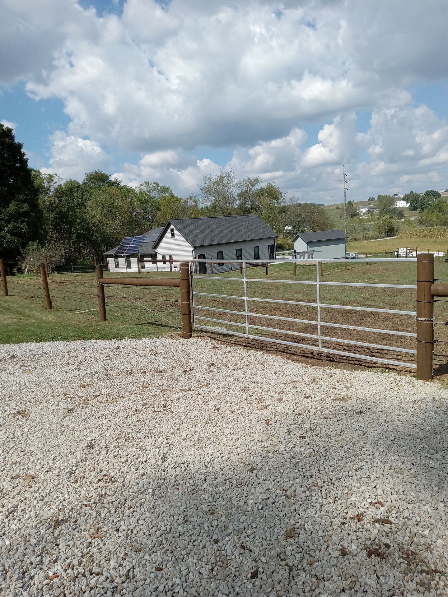 Gravel driveway leading to a fenced pasture, with a white barn under a partly cloudy sky.