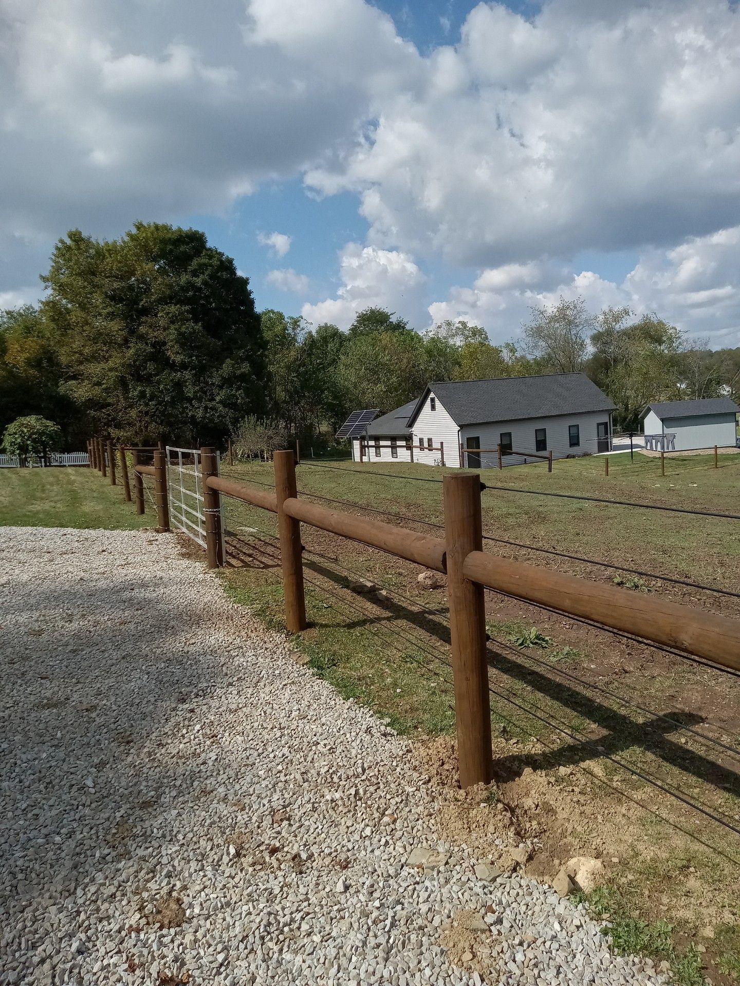 A gravel driveway leads to a wooden fence enclosing a grassy field with a white house in the background under a blue sky.