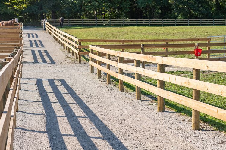 A wooden fence surrounds a dirt path leading to a horse stable.