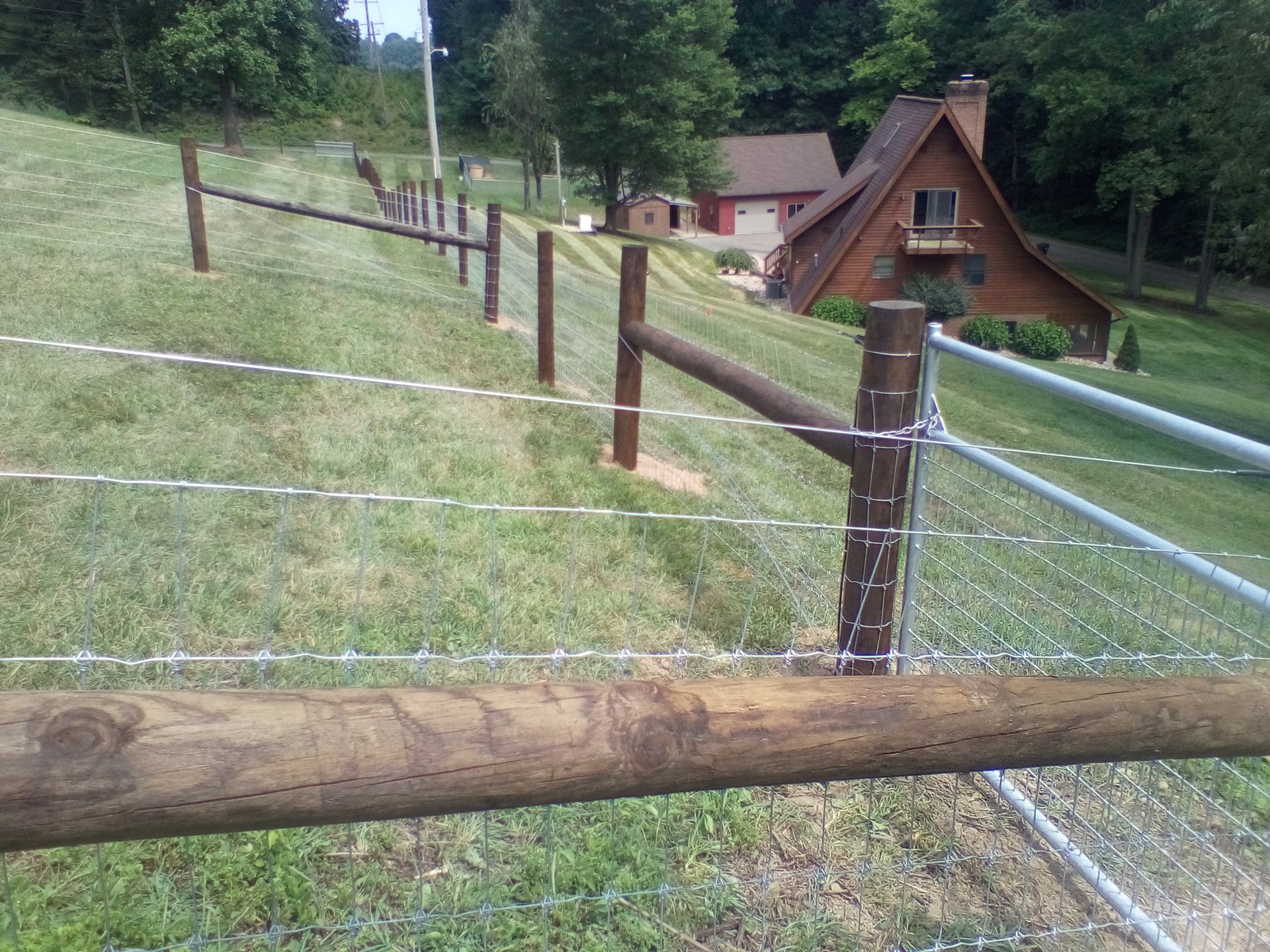 Brown wooden fence with barbed wire and metal mesh on a grassy hillside, near a house.