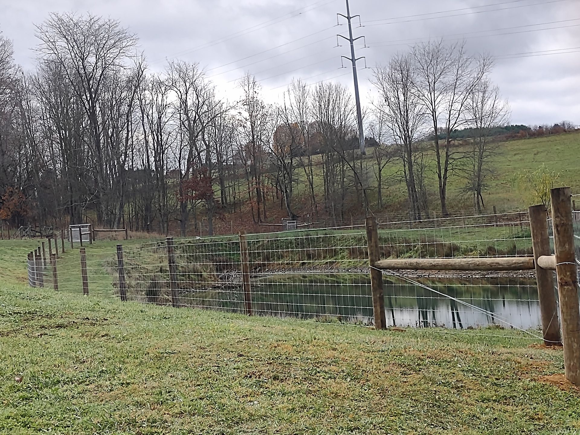 A wooden fence surrounds a pond in a field with trees in the background.