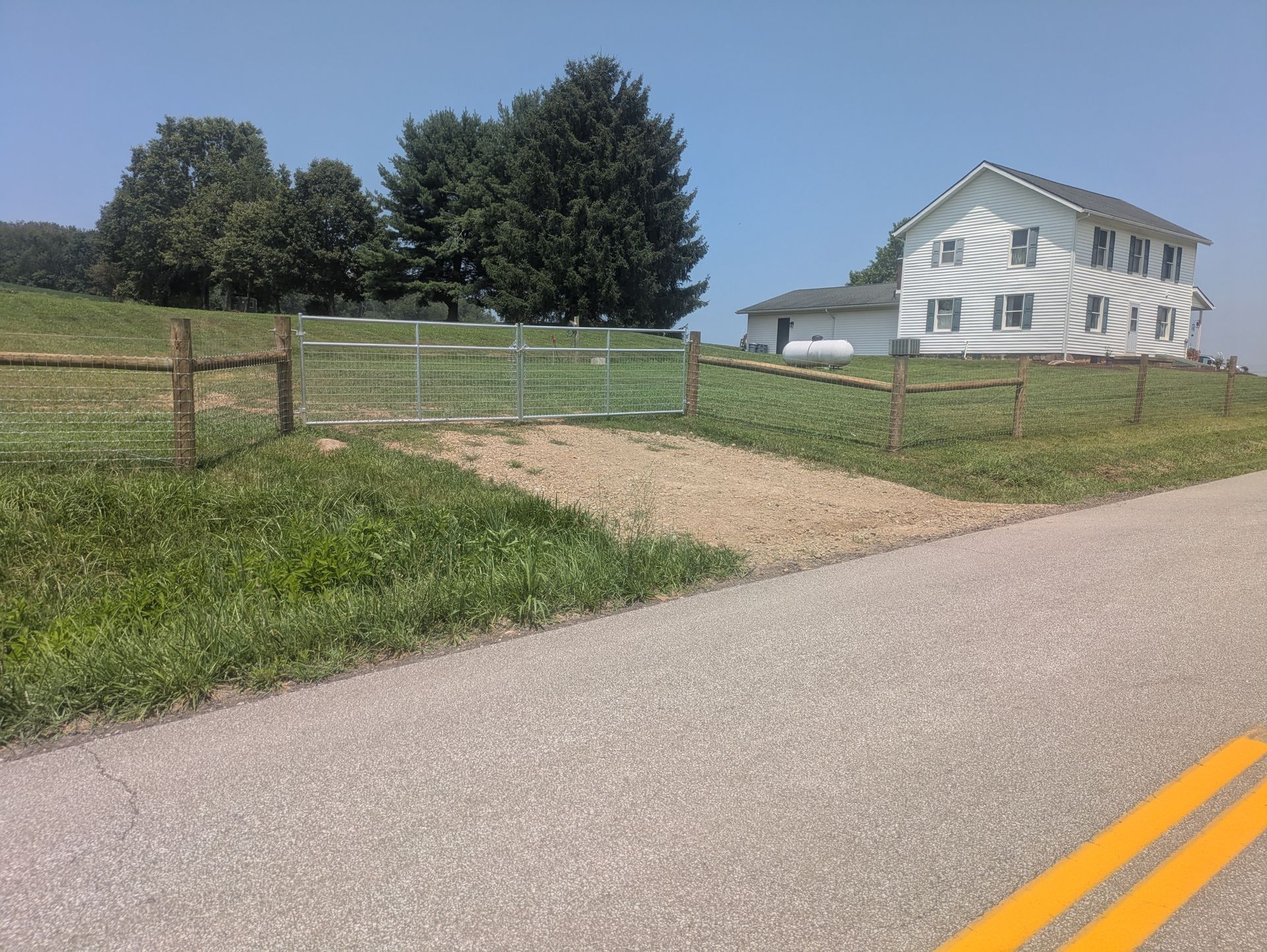 Rural scene: white house on a hill, gravel driveway, wooden fence, metal gate, road with yellow lines, blue sky, trees.