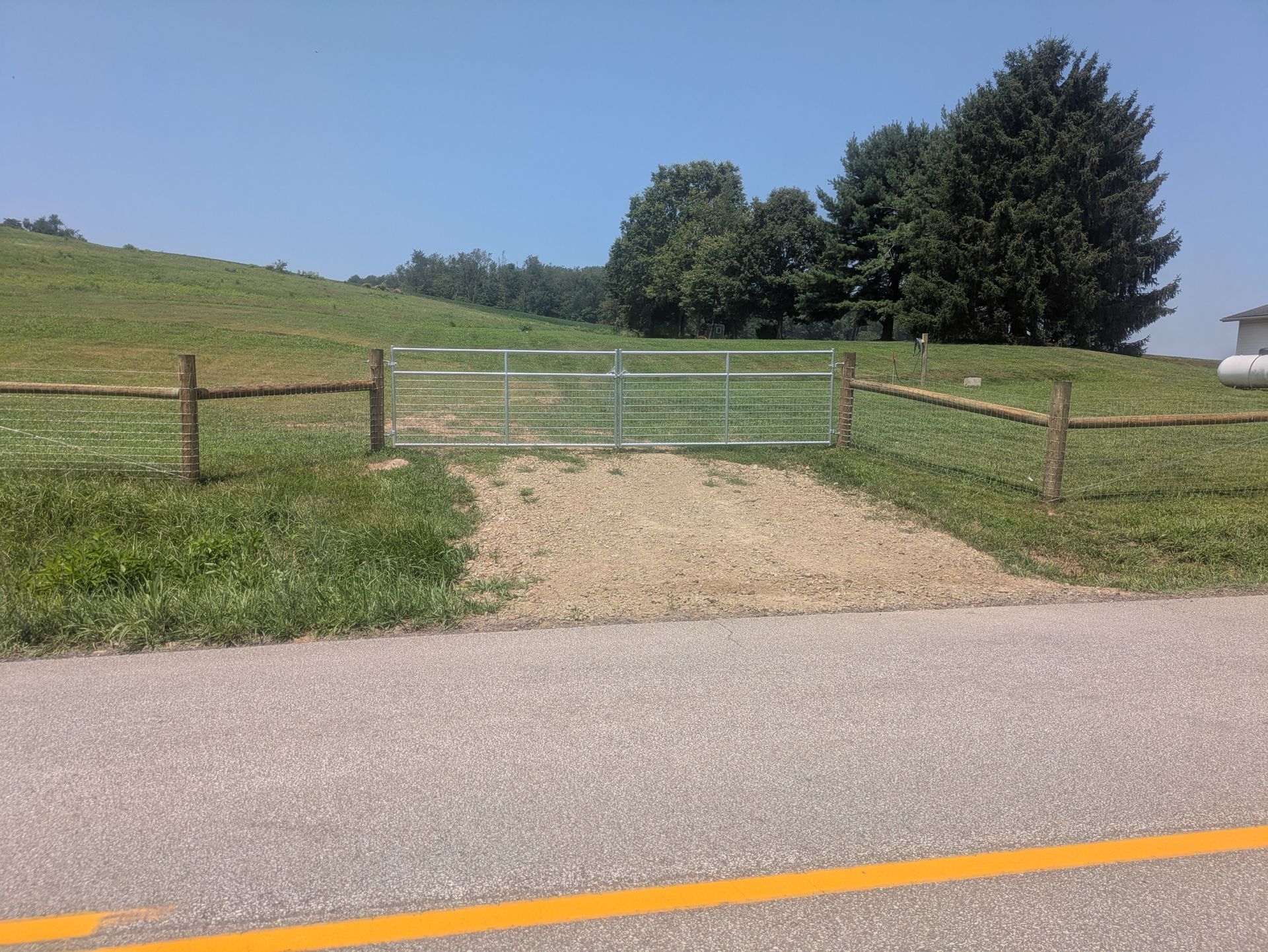 Metal gate in a rural setting, flanked by wooden posts and fencing, gravel path leading from a road.