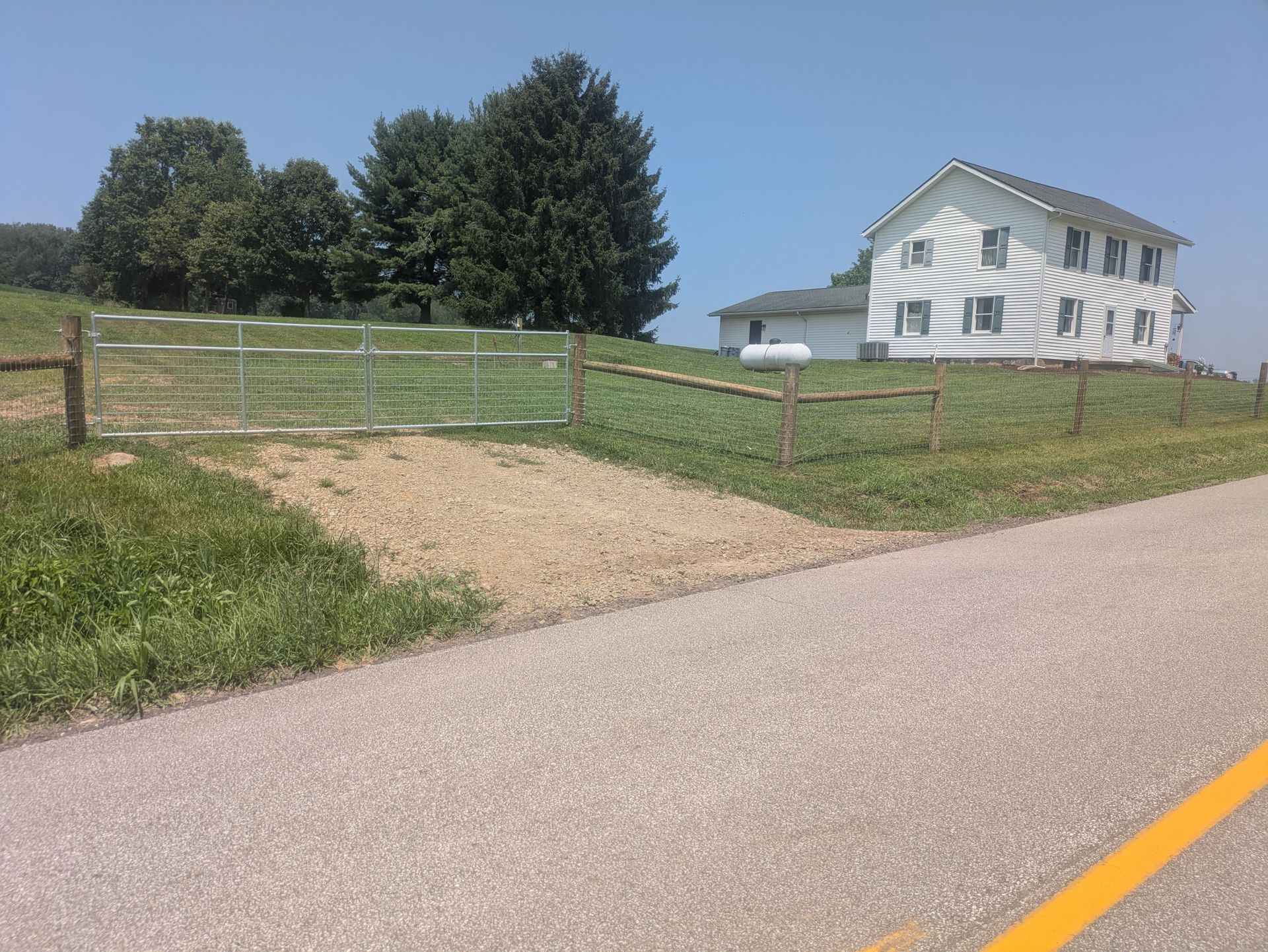 Gravel road leads to a house on a grassy hill; a metal gate and trees are in the foreground.