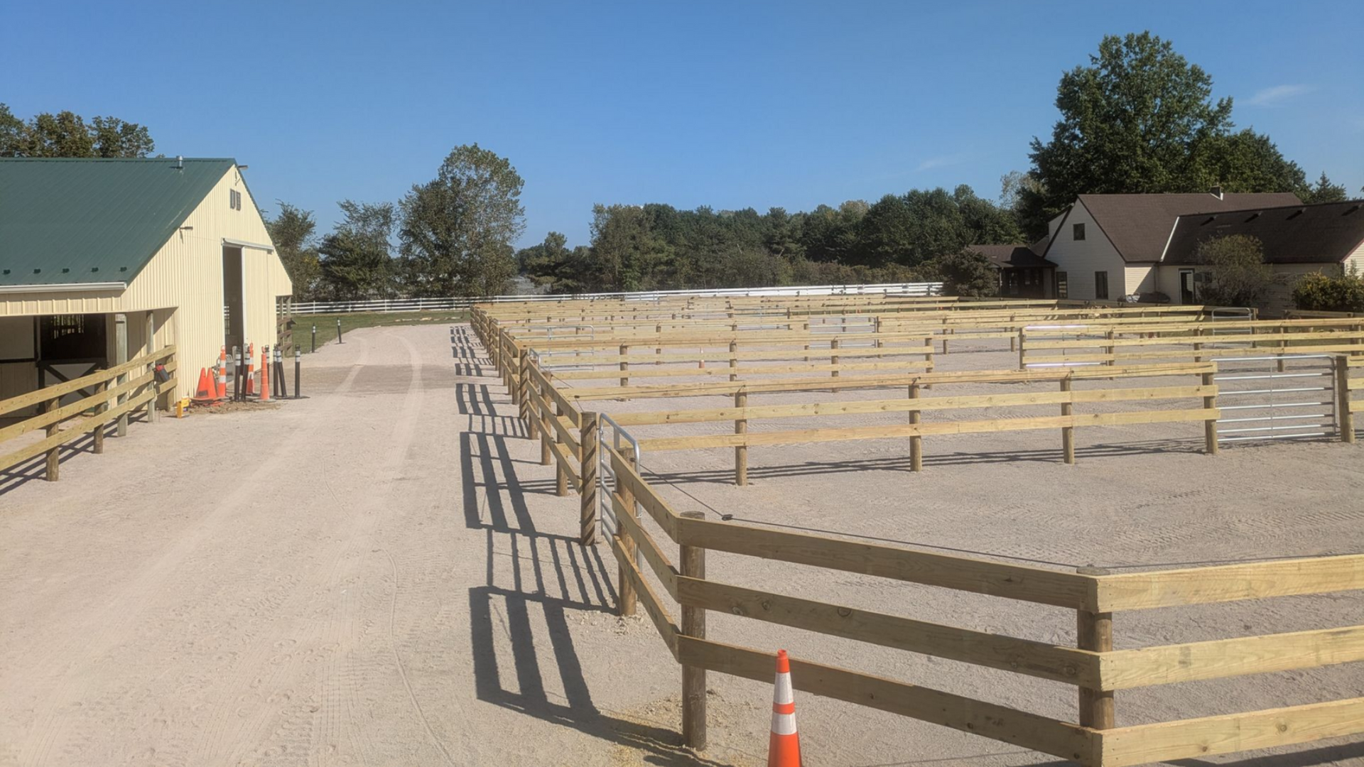 Gravel driveway lined with wood fences leading to open fields on a sunny day. Buildings in the background.