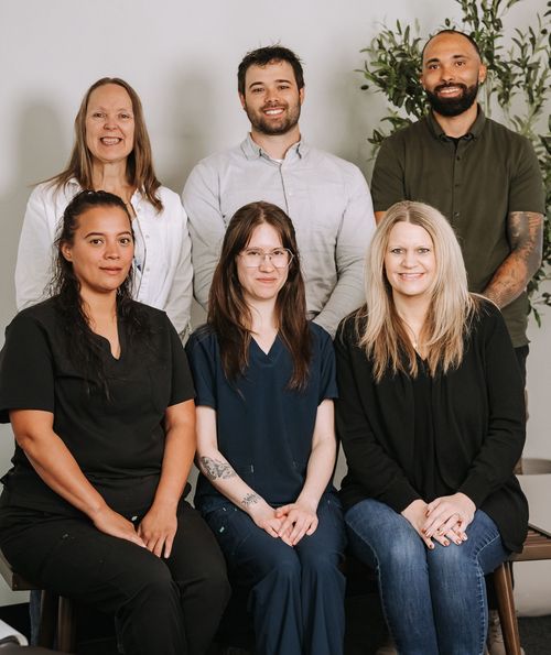 Five people seated and standing indoors, smiling in front of a light wall and plants.