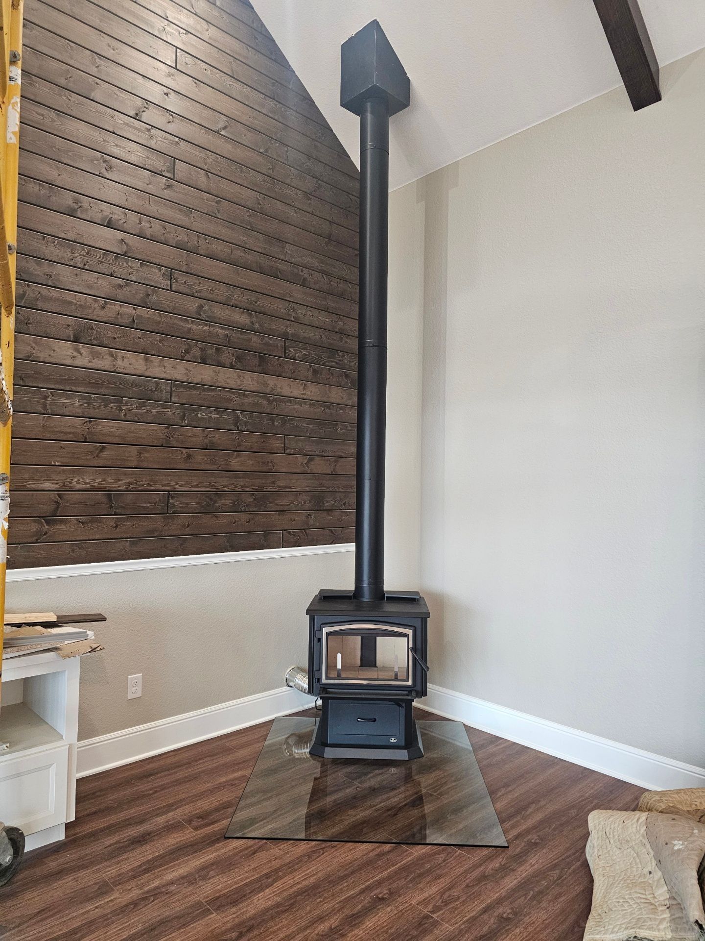 A wood-burning stove with black chimney pipe, on a glass hearth, in the corner of a room with wood paneling and light walls.