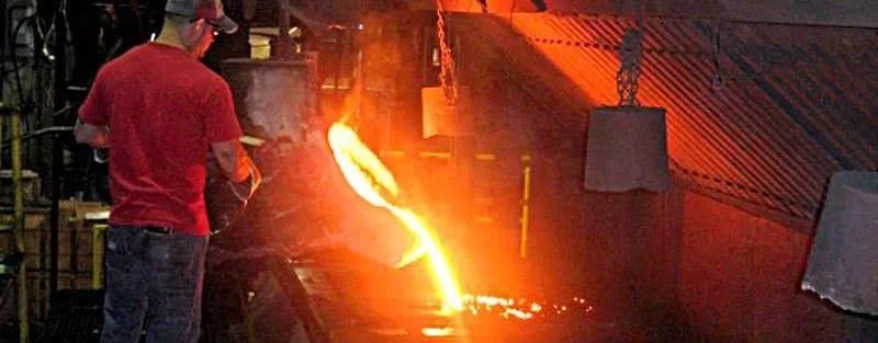 A worker pouring molten metal from a container in a factory. Orange glow, red shirt, and protective gear visible.