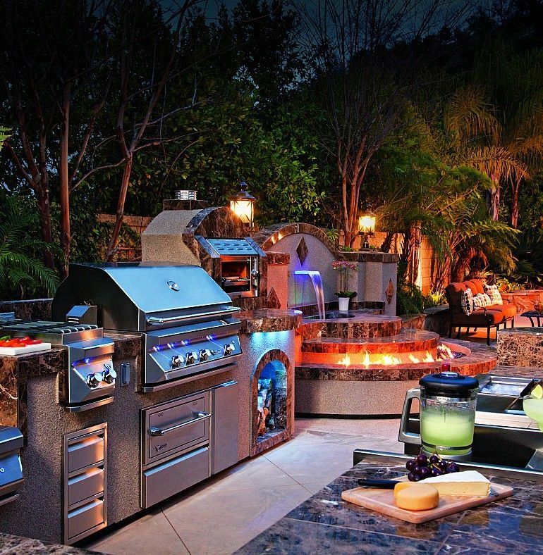 Outdoor kitchen with grill, pizza oven, and a water feature, illuminated at night.