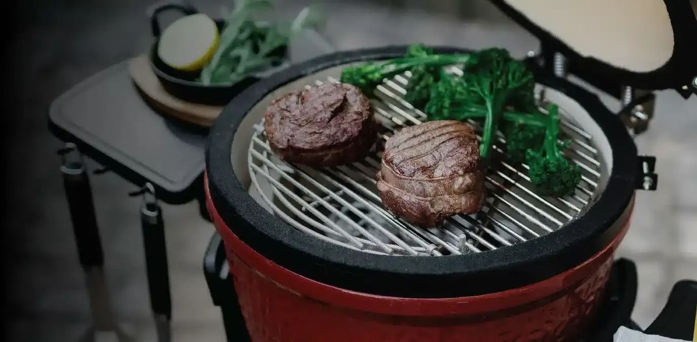 Red grill with steaks and greens cooking on a metal grate. Side table with lemon and arugula.