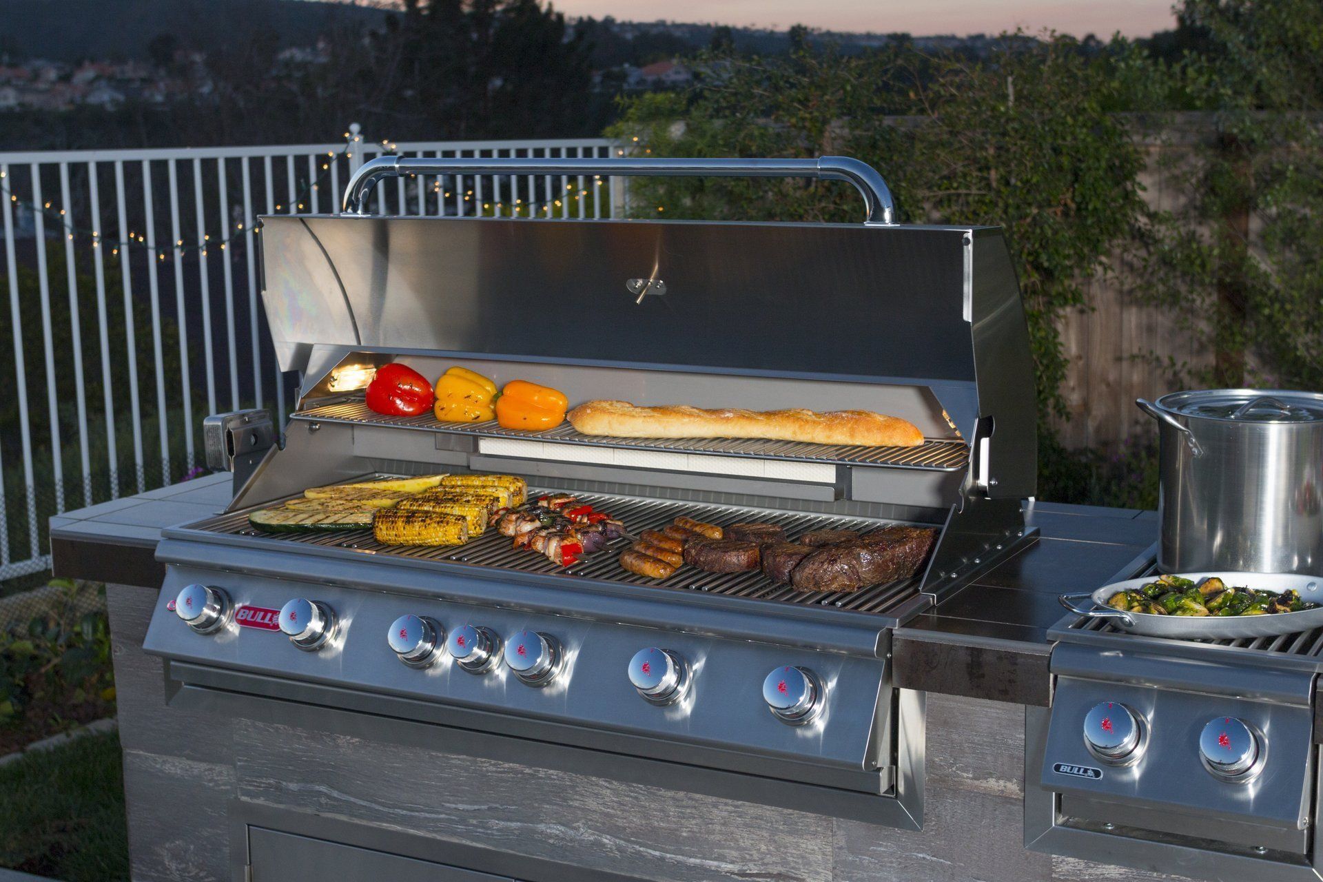 Outdoor grill with various foods cooking on the grates, stone countertop.