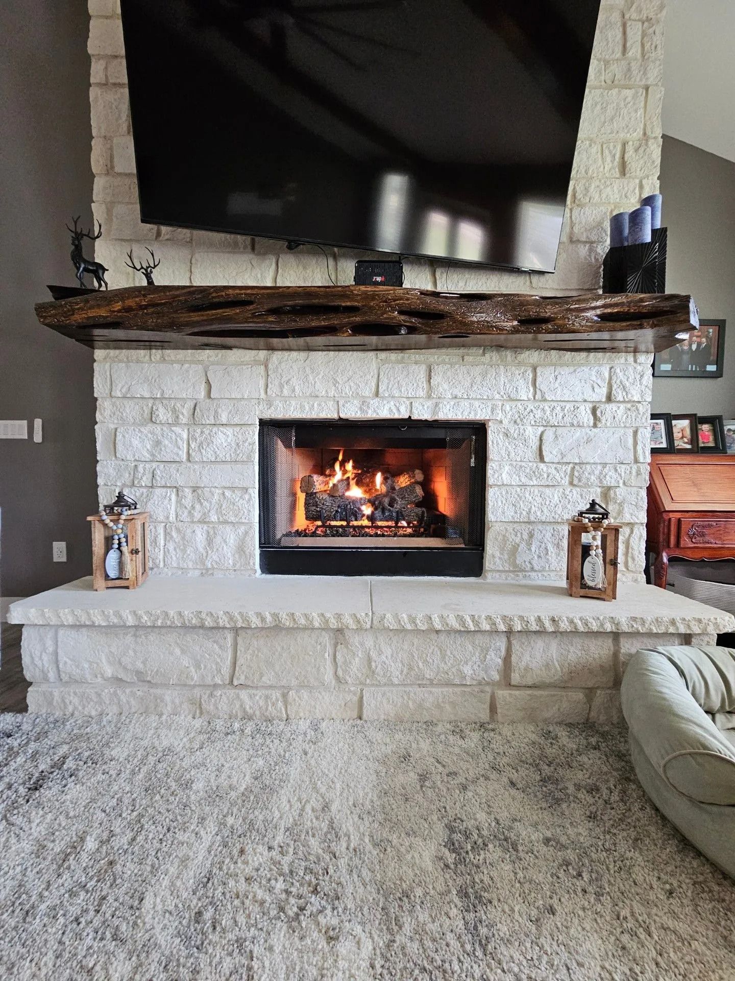 Stone fireplace with burning fire, large TV, and rustic wooden mantel. Two lanterns sit on the hearth.