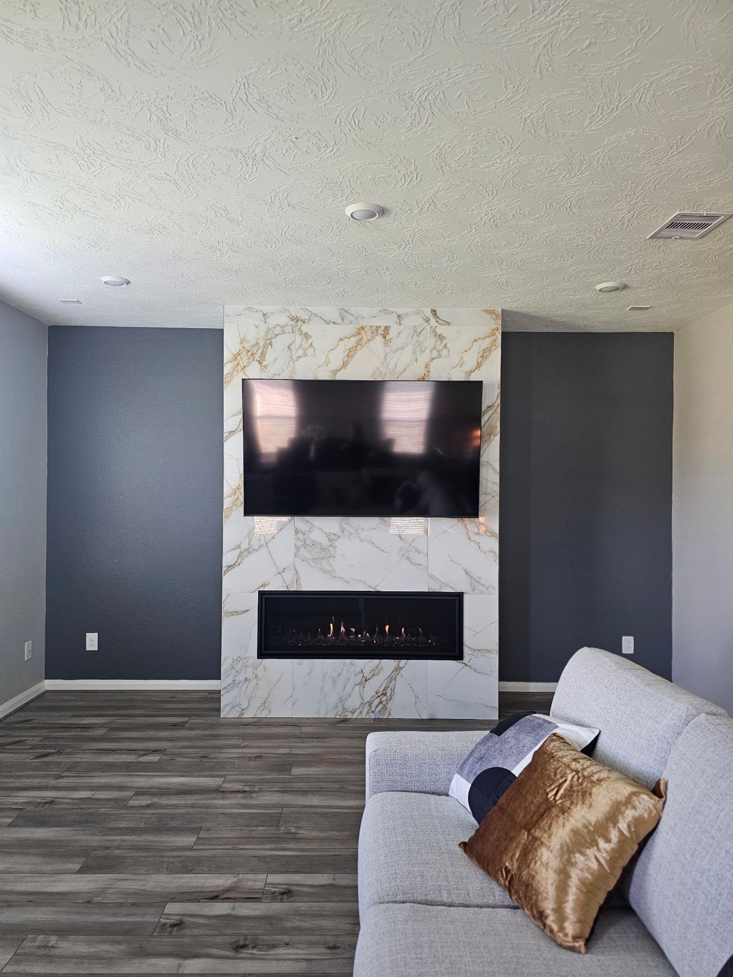 Living room with a fireplace and TV mounted on marble. Gray walls, wood-look floor, and a gray sofa.
