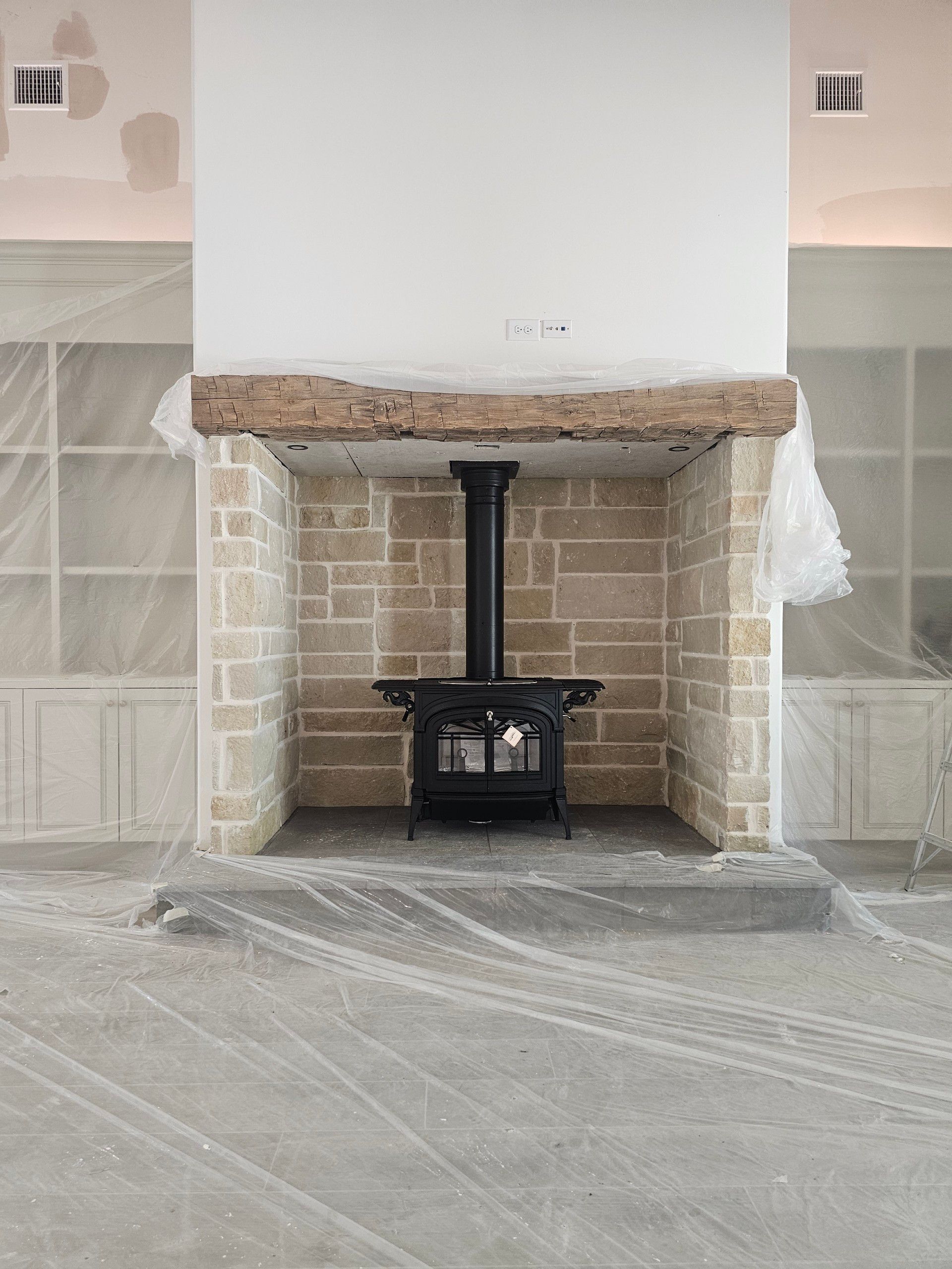 Black wood-burning stove inside a stone fireplace with a wooden beam mantel. Under construction, unfinished room.