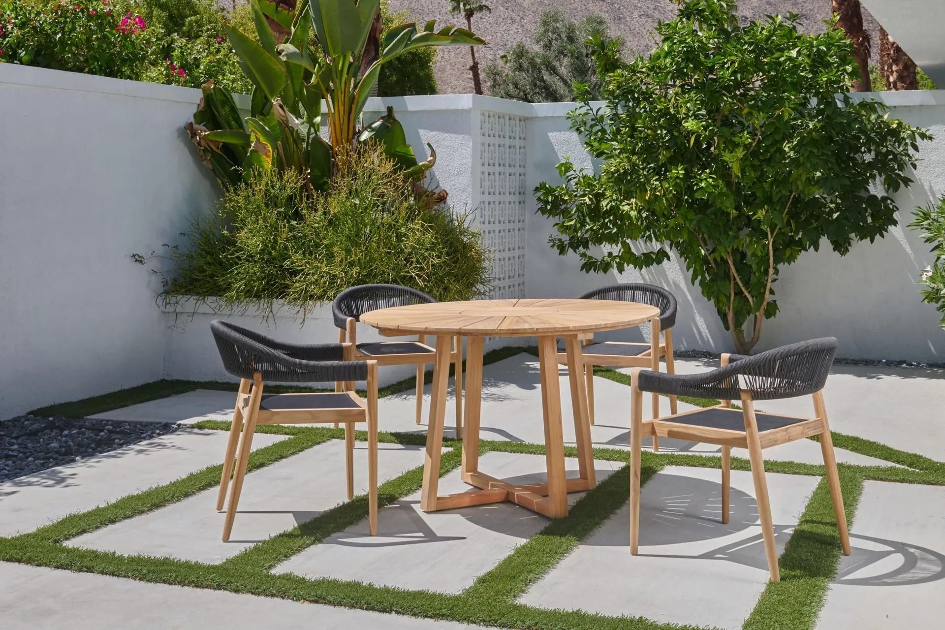 Outdoor dining set: round wooden table, four chairs with woven backs, on a patio with greenery.
