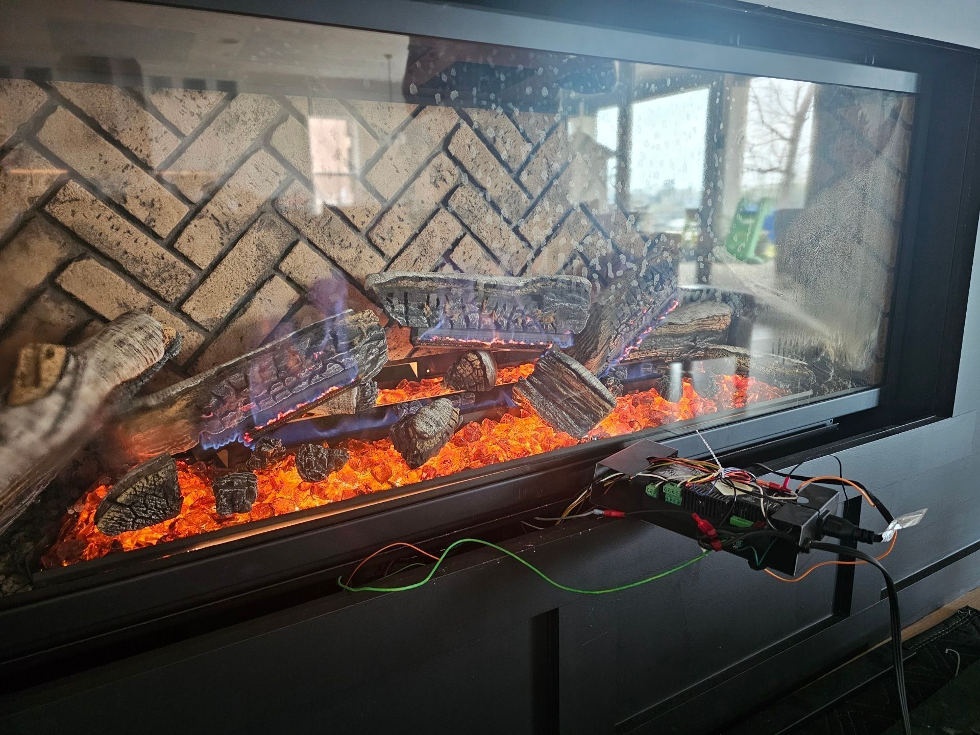 Black electronic device near a lit fireplace with decorative faux logs, against a brick background.