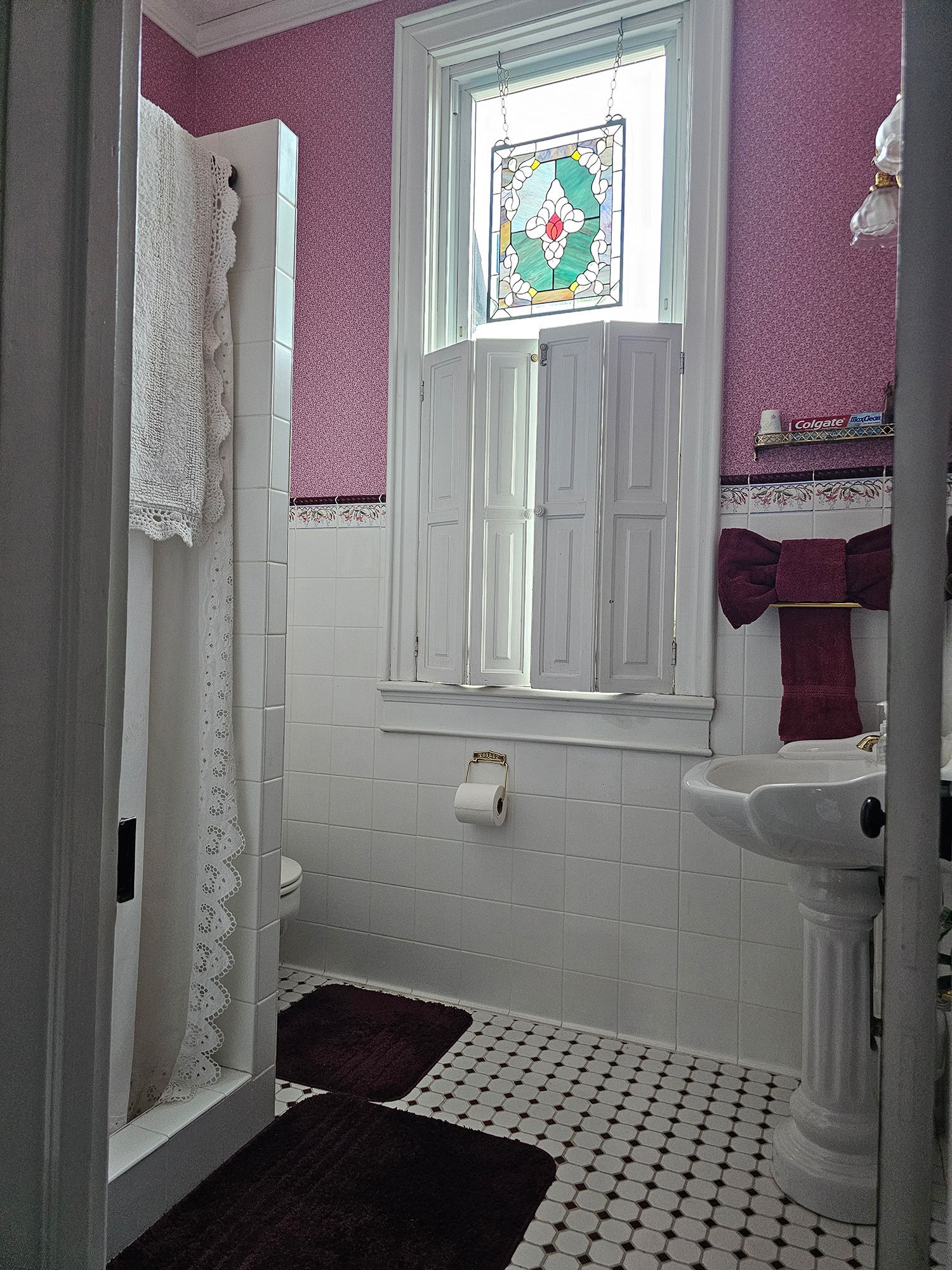 A vintage-style bathroom with a stained-glass window, pink wallpaper, white tile, a pedestal sink, and a lace shower curtain.