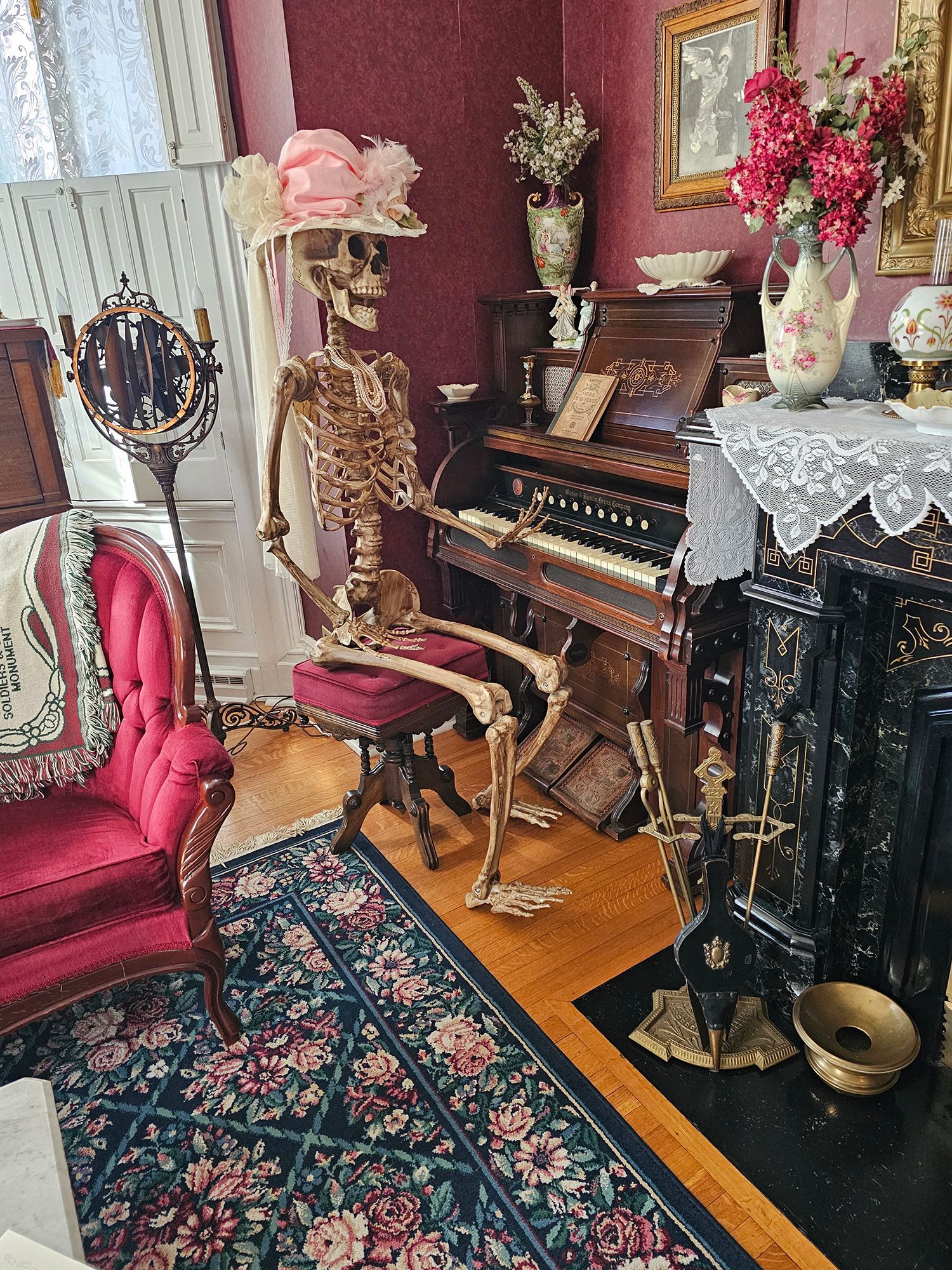Skeleton wearing a hat, playing an organ in a Victorian-style room. Room has red walls, antique furniture, and a rug.