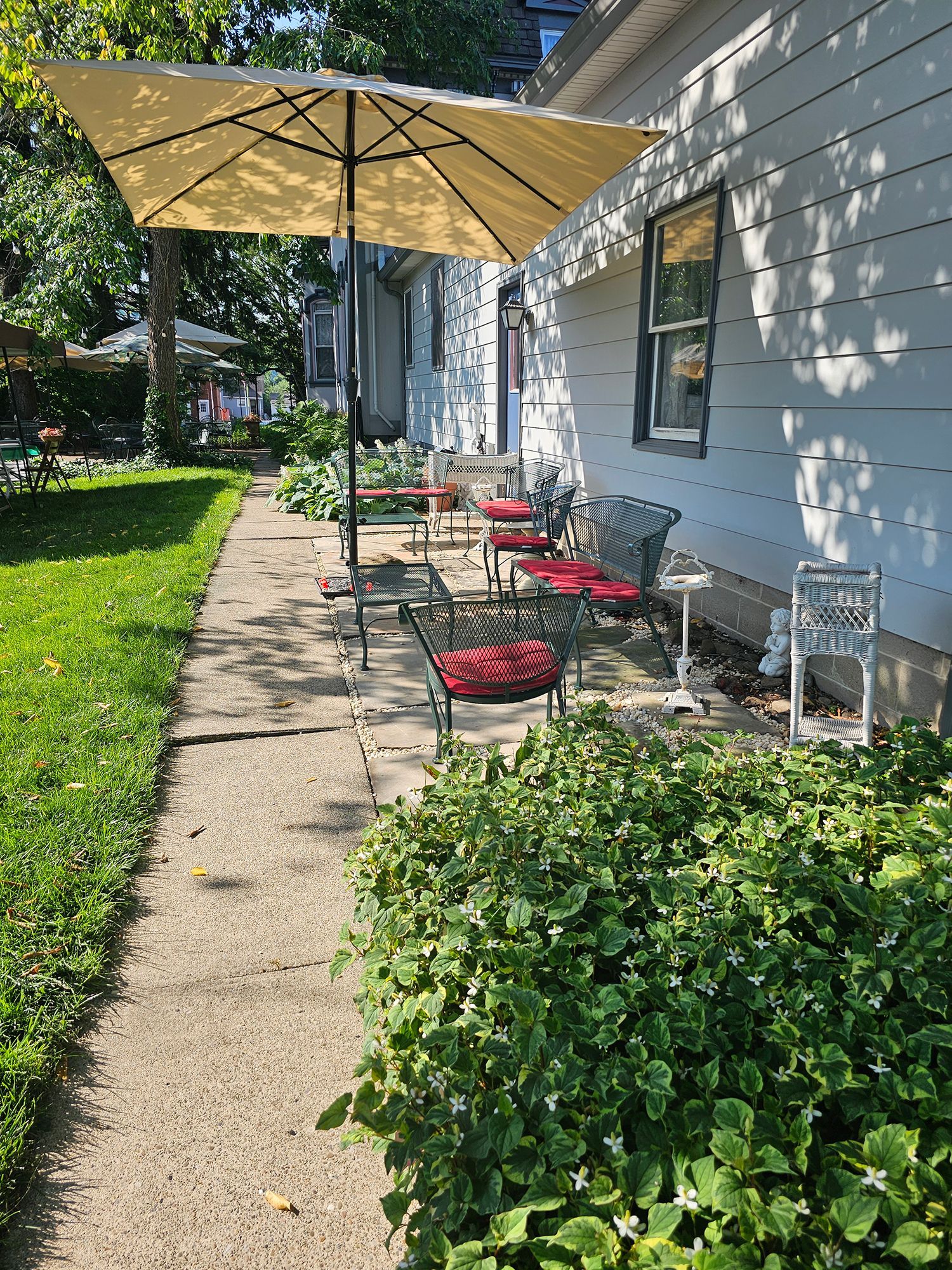 A concrete path leads past a house with outdoor seating under a large yellow umbrella. Green grass and plants border the path.