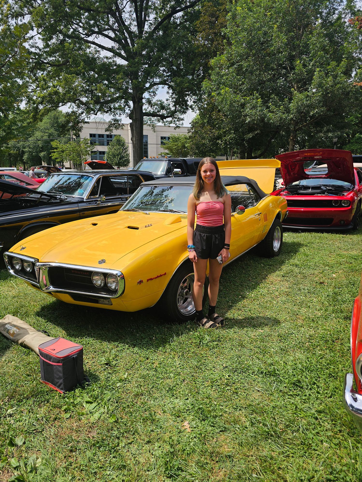 A young woman stands in front of a bright yellow convertible car with its hood open at an outdoor car show.