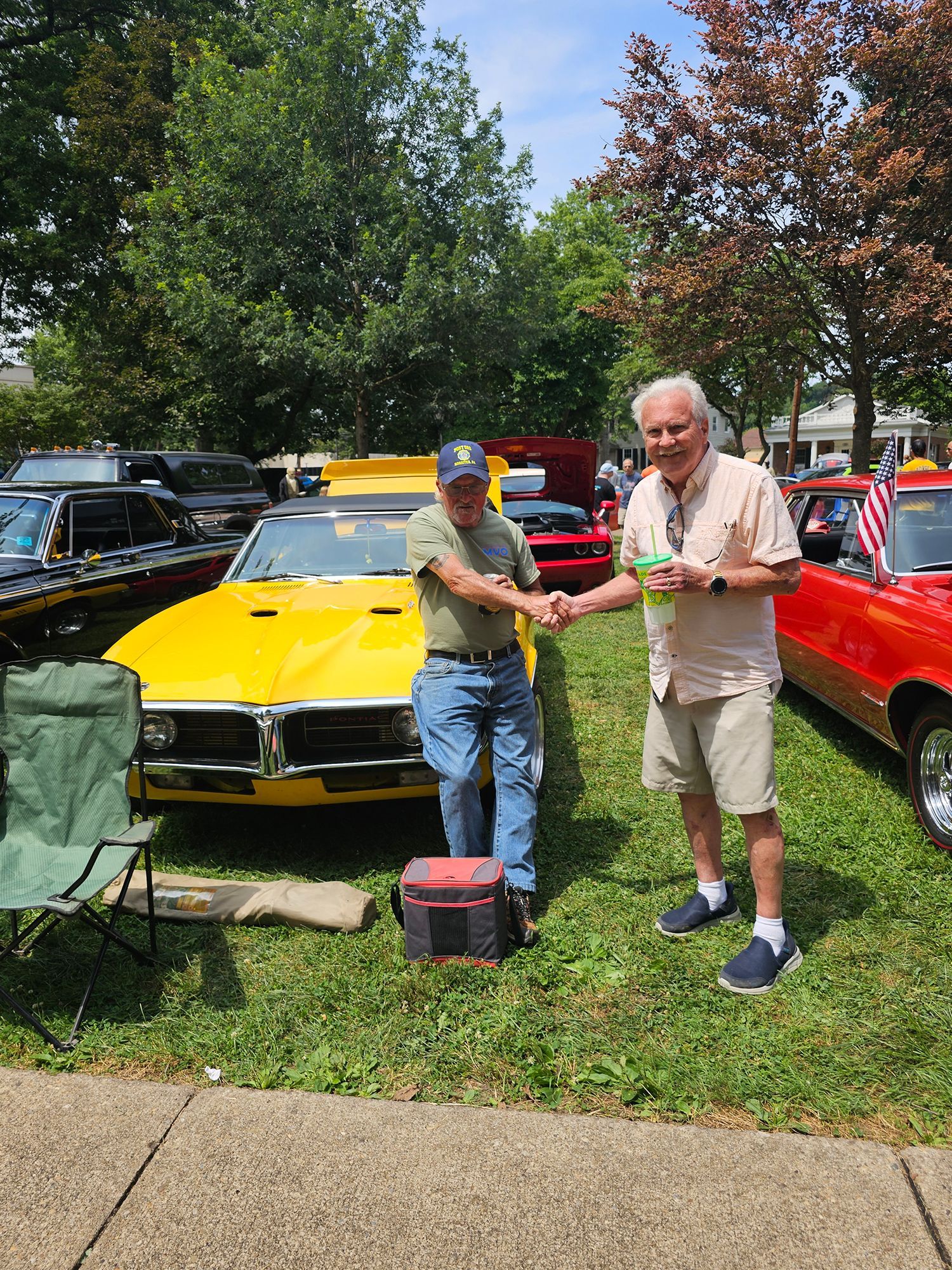 Two men shaking hands next to a yellow classic car at an outdoor car show. Green grass and other cars are visible.