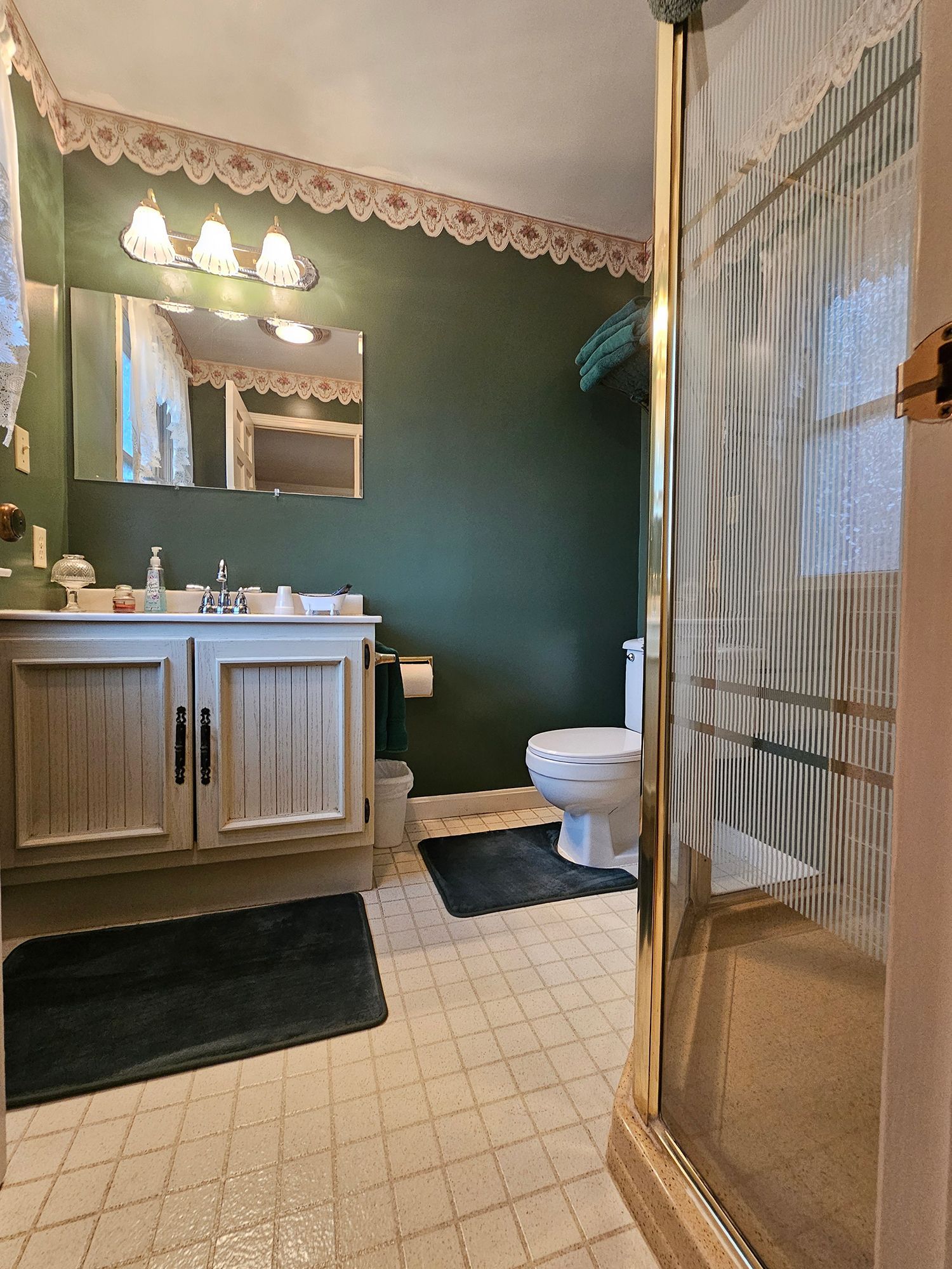 Bathroom with a light-colored vanity, toilet, and shower. Dark green wall, lace trim, and a patterned tile floor.