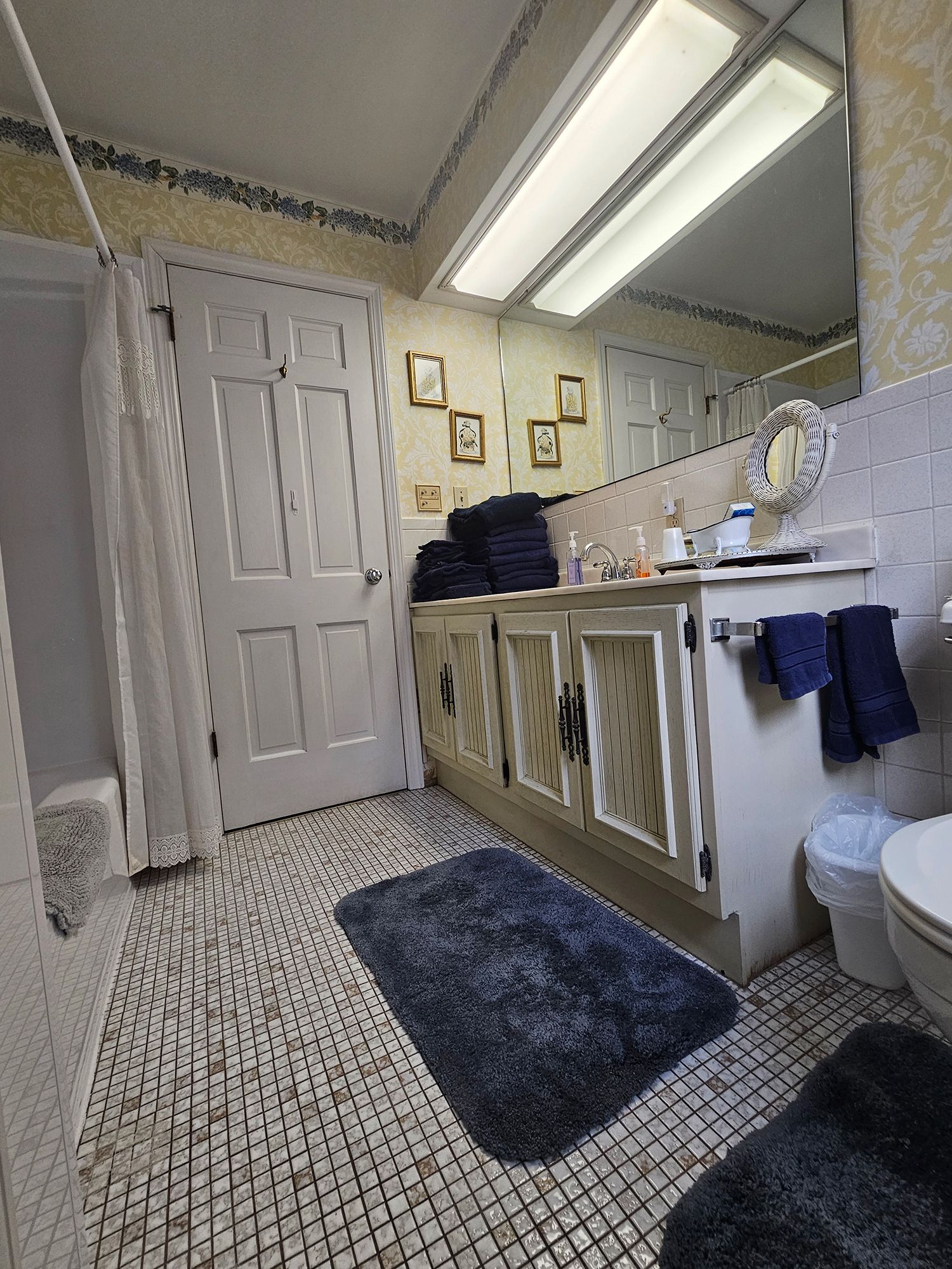 Bathroom with white cabinets, patterned tile floor, and a blue rug. A shower and a door are visible.