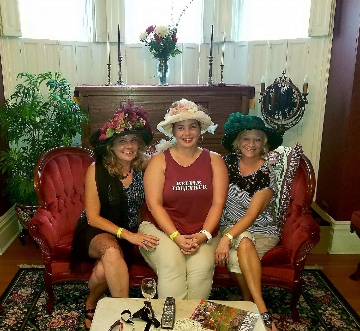 Three women in decorated hats pose on a red Victorian sofa. They smile, with a table of items in front.