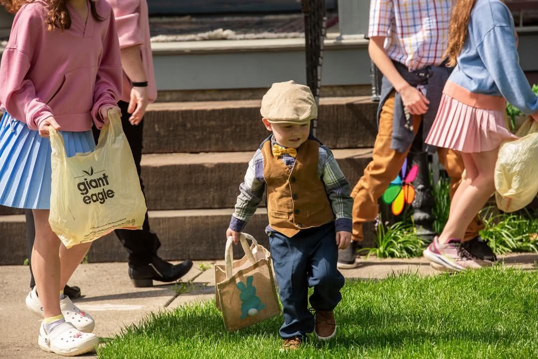 A young boy in a cap carries a bag, followed by others holding bags, walking on grass in front of a building.