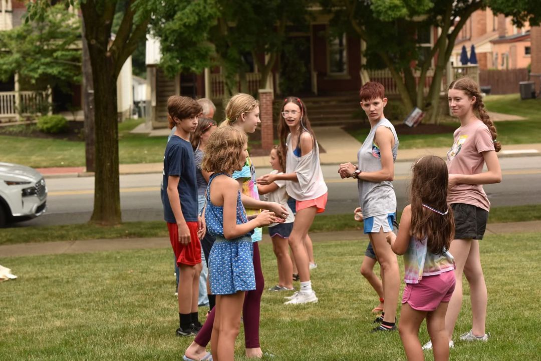 A group of children in casual clothes play outside on a grassy lawn, smiling and interacting near a street.